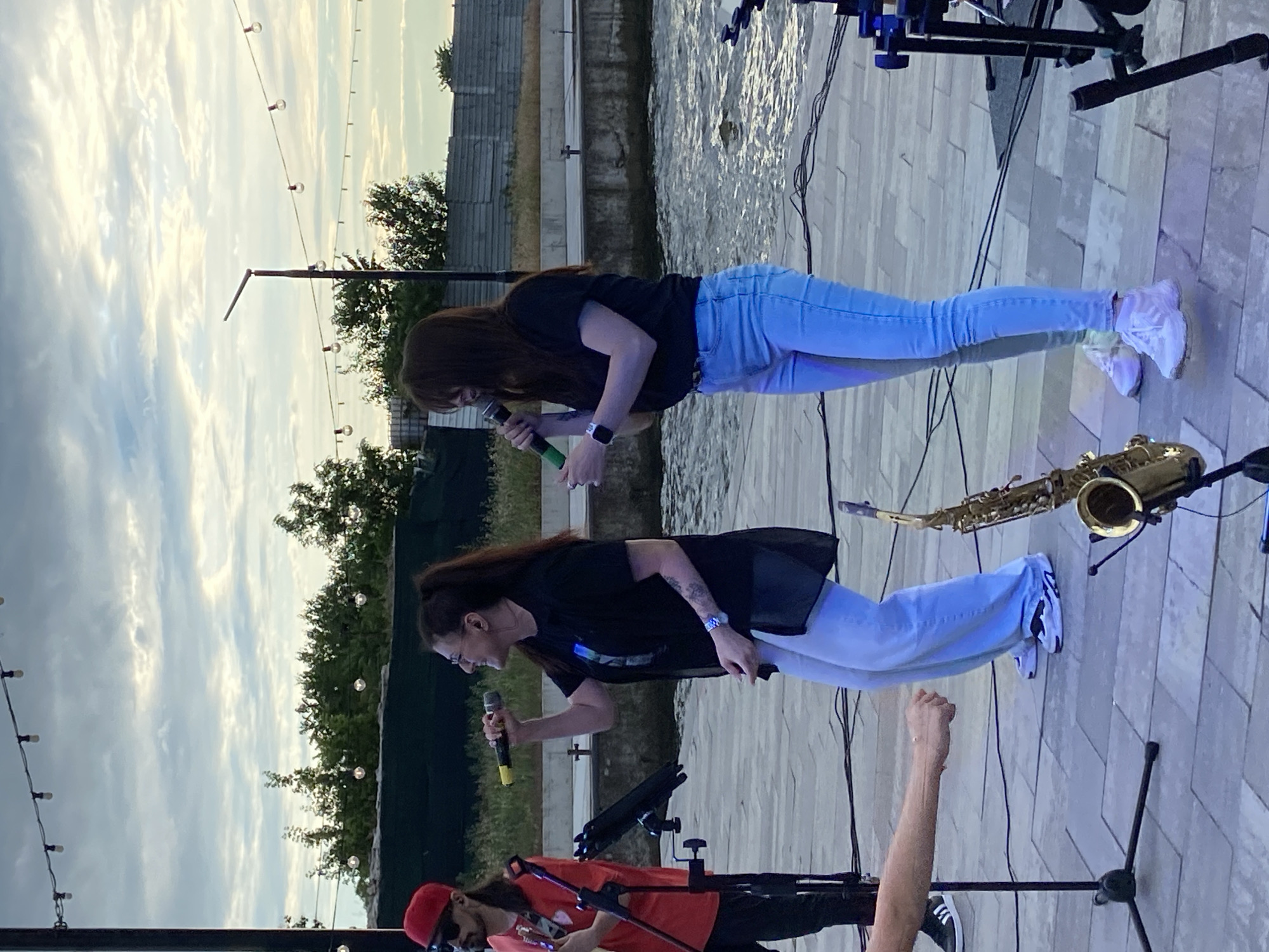 Two women are singing into microphones on an outdoor stage. The woman on the left has her hair in a ponytail and is wearing a black shirt and light-colored pants. The woman on the right has long, dark hair and is wearing a black t-shirt and light-colored jeans. A saxophone is visible on the stage in front of the woman on the right. In the background, there is a body of water and trees. String lights are hanging overhead. The scene appears to be at dusk.