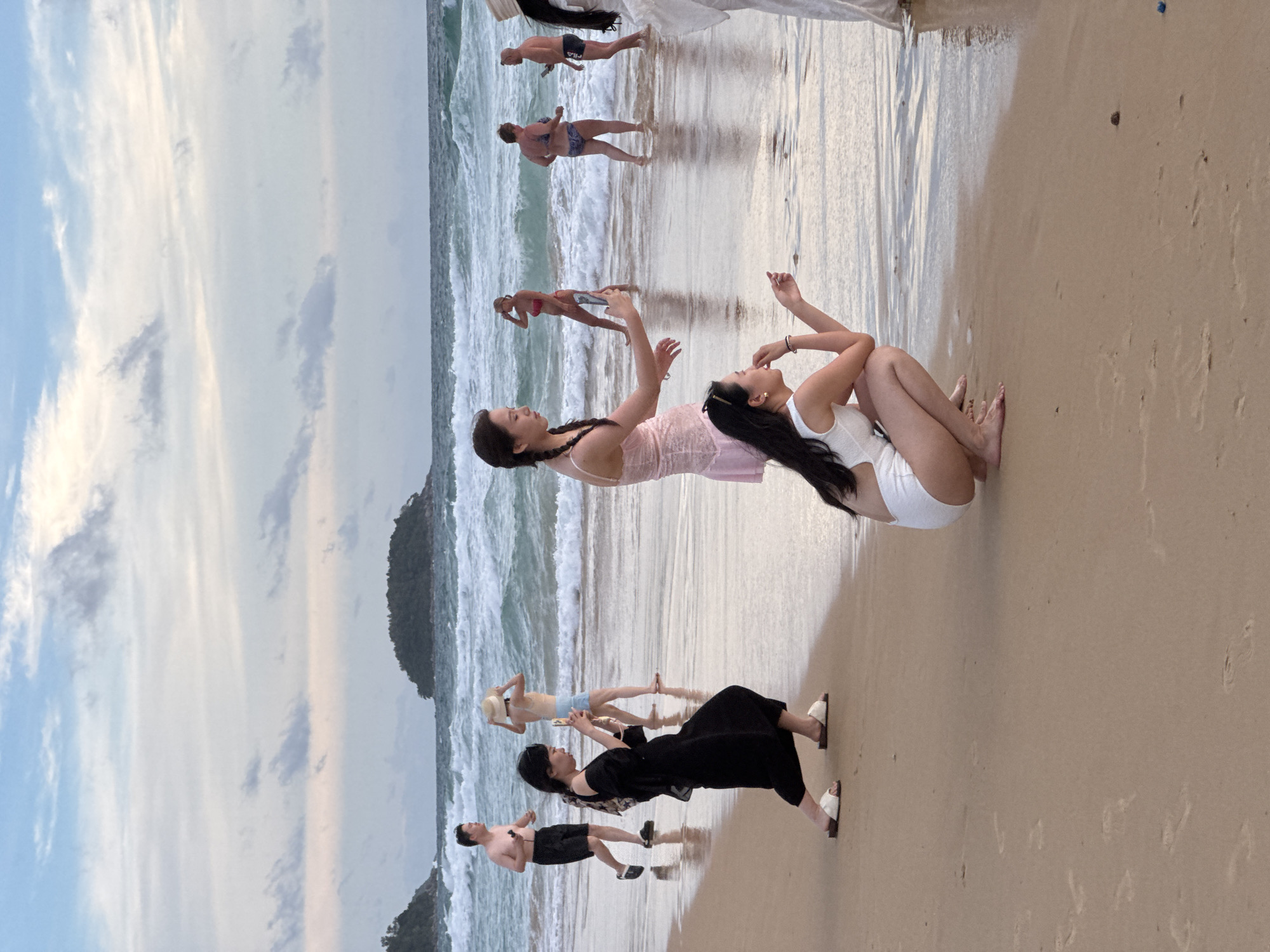 In Karon, Thailand, a group of people are on a sandy beach with the ocean in the background. Several individuals are in the water, while others are on the shore. The sky is overcast with clouds. In the foreground, a woman in a black dress is squatting and holding a smartphone, seemingly taking a photo. Another woman in a white swimsuit is also squatting nearby. Further back, a man in swim trunks is walking, and a woman in a straw hat is standing. Other individuals are visible in the distance, wading in the ocean. The waves are moderate, and the tide appears to be low. Islands are visible in the distance. The sand is damp from the receding tide, showing footprints.