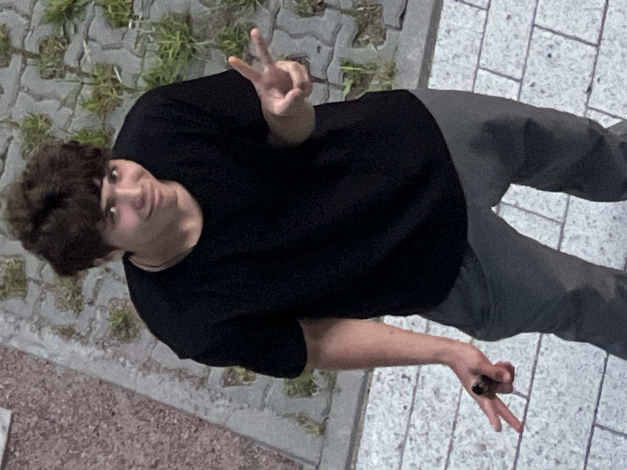 A young male subject is depicted standing outdoors in the city of Unknown, Russia, viewed from a slightly elevated perspective. The individual possesses dark, curly hair and fair skin. He is dressed in a black short-sleeved t-shirt and dark grey trousers. A light-colored necklace or chain is visible around his neck. The subject is looking towards the camera with a subtle smirk. His right hand (viewer's left) is raised, forming a "peace" sign with the index and middle fingers extended. His left hand (viewer's right) is also raised, forming a "peace" sign, and he is holding a dark, cylindrical object, consistent with a cigar, lodged between his extended index and middle fingers. The ground beneath the subject is paved with light-colored, possibly white or grey, square or rectangular stones, showing scattered dark specks. To the immediate left of the subject, the ground transitions to a reddish-brown, textured surface. Behind the subject, a grey vertical surface or wall, constructed of irregularly shaped blocks or stones, features small green plants growing from its crevices. The scene is illuminated by uniform ambient light.