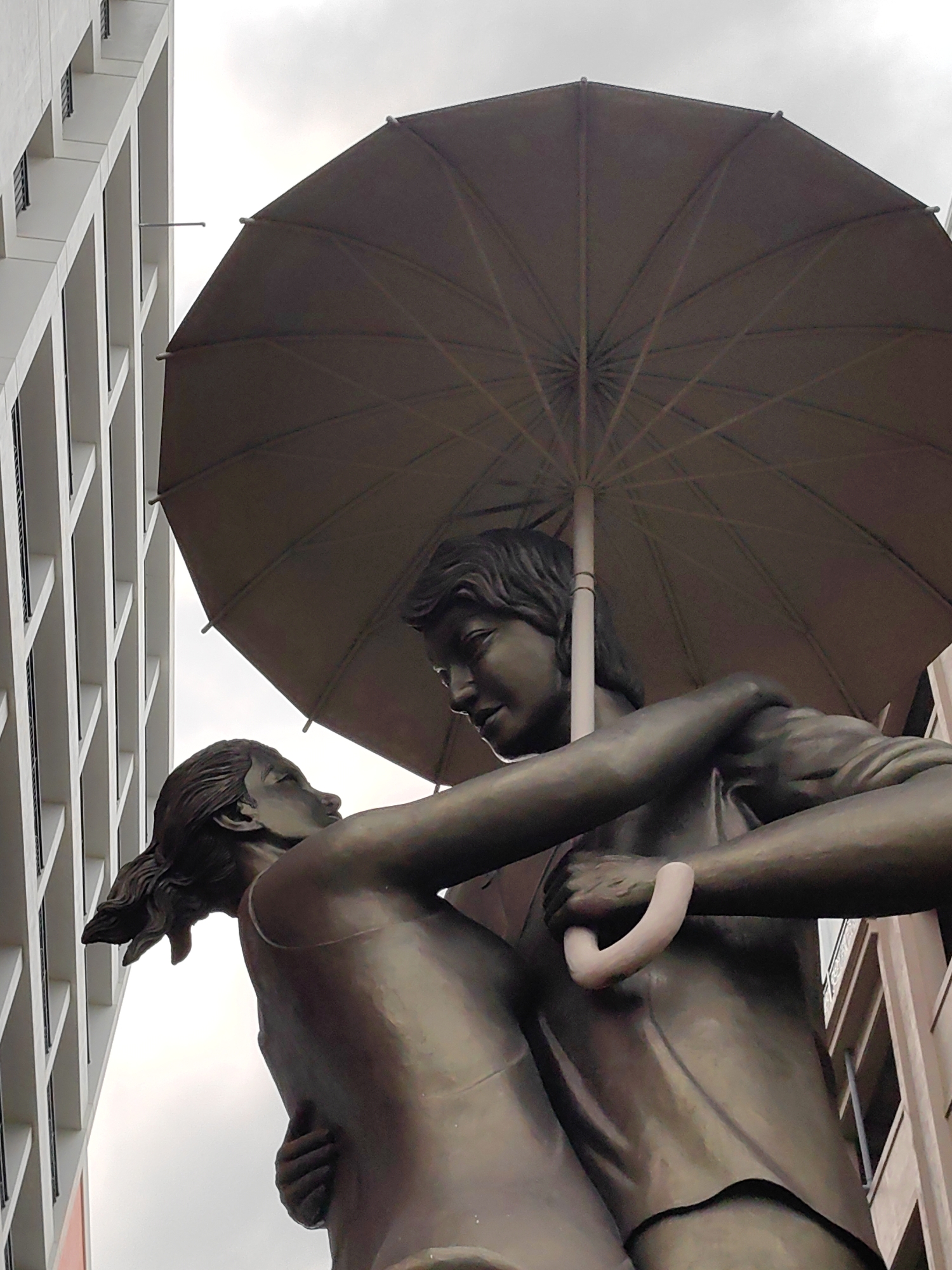 A bronze statue of a couple embracing under an umbrella. The statue is set against a backdrop of modern buildings, one white and one pink. The sky is overcast. The location is Tuan Chau Ward, Vietnam.