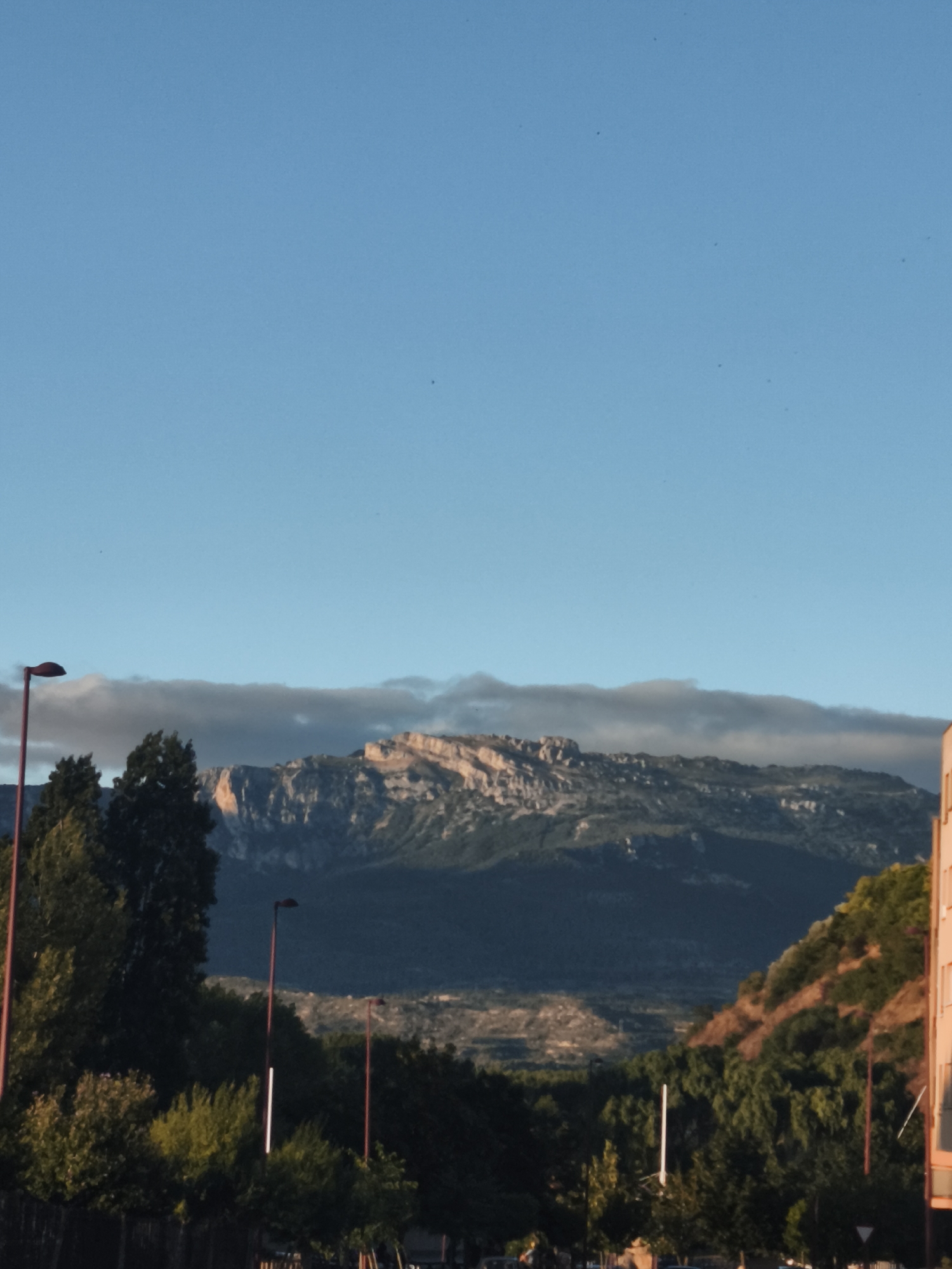 A vertical, eye-level, outdoor shot shows a mountainous landscape in the distance, under a clear blue sky with a few faint, wispy clouds. In the foreground, there are trees and two streetlights with red poles. A building with an orange facade is visible on the right side of the frame. The image appears to be taken in the city of Haro, Spain.