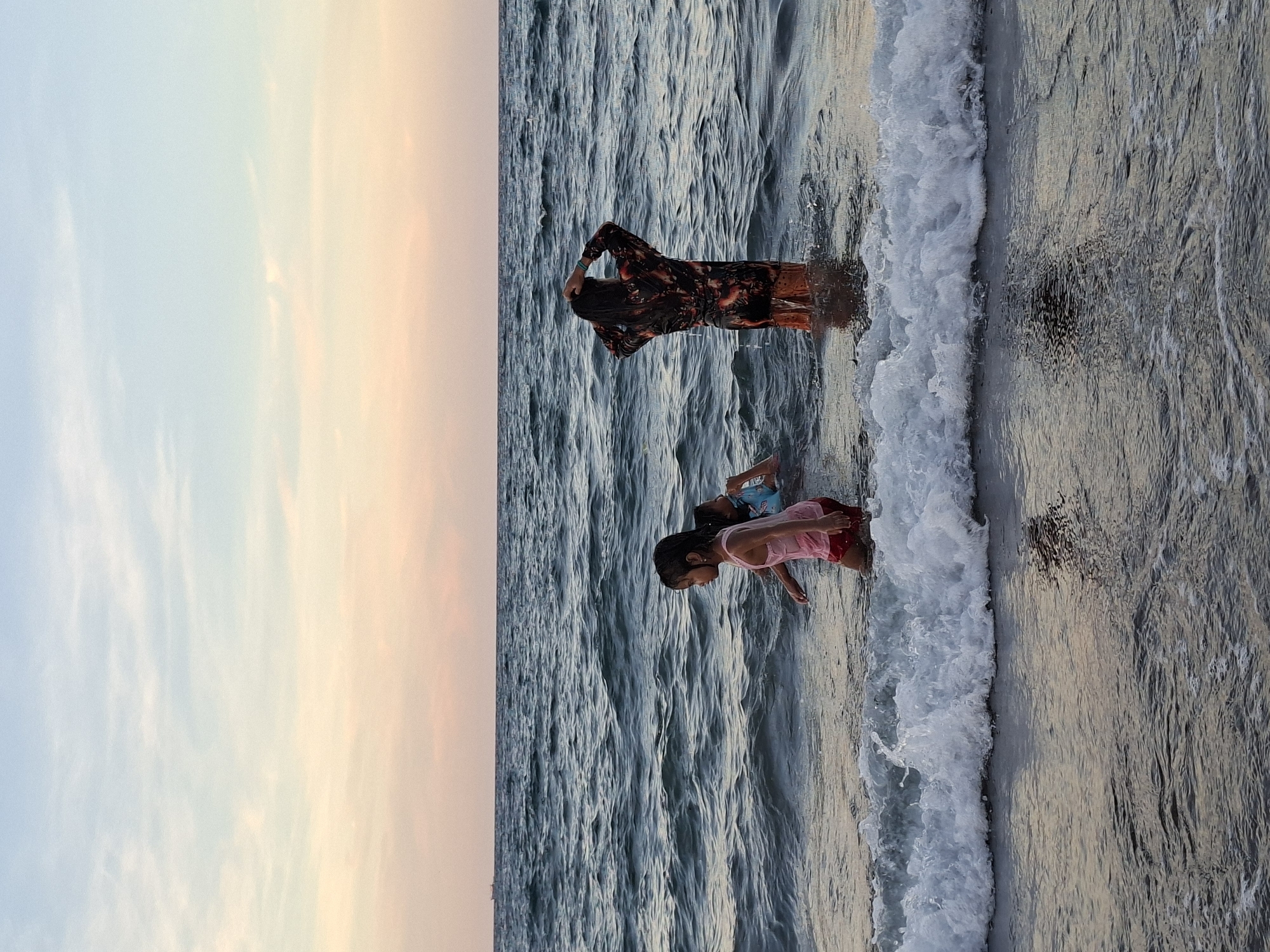 A low-angle, landscape-oriented shot depicts a beach scene at dusk. In the background to the left, the sky transitions from pale blue to peach hues. The ocean dominates the majority of the scene, showing wave patterns. Two children, likely siblings, are playing by the shore, with a woman in the background standing in the water near the incoming waves. The location is presumably along the coast of Lhokseumawe, Indonesia, given the instructions, but the image offers no specific landmarks to confirm it.