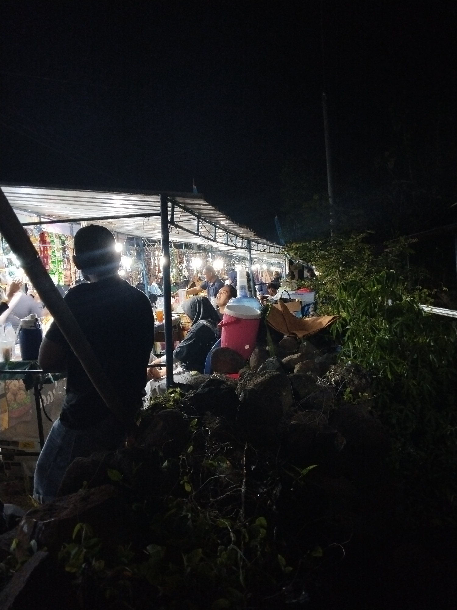 This nighttime image captures an outdoor market or street food area, likely in Jenggrik, Indonesia. In the immediate foreground, a person is seen from behind, wearing a dark t-shirt and light-colored trousers, standing near a wooden or metal pole. Their head is silhouetted against the bright artificial lights emanating from the stalls behind them.
Beyond this foreground figure, a series of temporary market stalls are brightly illuminated by fluorescent lights under simple canopies, possibly made of corrugated metal. These stalls appear to be selling various items, with numerous packets of goods visible hanging from the structures. Several people are gathered among the stalls, including an individual wearing a dark hijab who is seated and looking towards the left. Another person, a man, is visible in the background engaged in conversation.
The right side of the image and the lower foreground are dominated by dark rocks and dense green vegetation, contrasting with the well-lit market area. A large blue and a red cooler are also visible on the ground near the rocks. The setting is clearly at night, with the darkness of the surroundings emphasizing the vibrant light of the market. The scene conveys a sense of community and activity, typical of a street market or food vendors operating after sundown. No clear, legible text is visible.