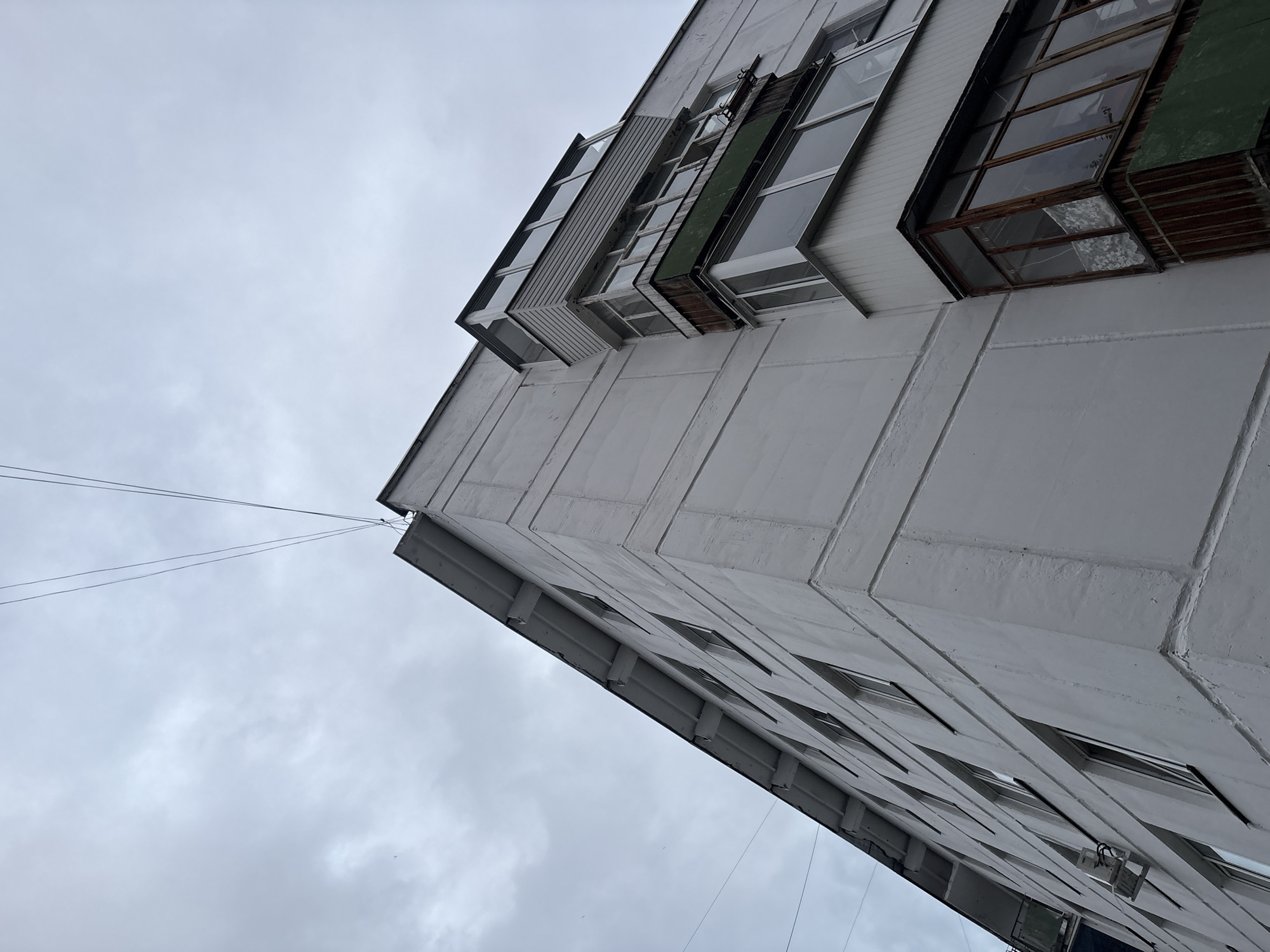 A low-angle outdoor shot captures the exterior of a stark, white, multi-story apartment building under a heavily overcast sky. The building's corner is prominently featured, with the facade featuring horizontal and vertical relief lines. Numerous windows are visible along the length of the building, some with dark frames. On the right side of the building, a balcony with enclosed glass panes and wooden trim juts out. Another, smaller balcony with lighter siding is situated above it. Power lines stretch diagonally across the frame from the top left to the upper right corner. An outdoor air conditioning unit is mounted on the wall of the building towards the bottom left. The overall impression is one of urban, somewhat weathered architecture under a gloomy sky.