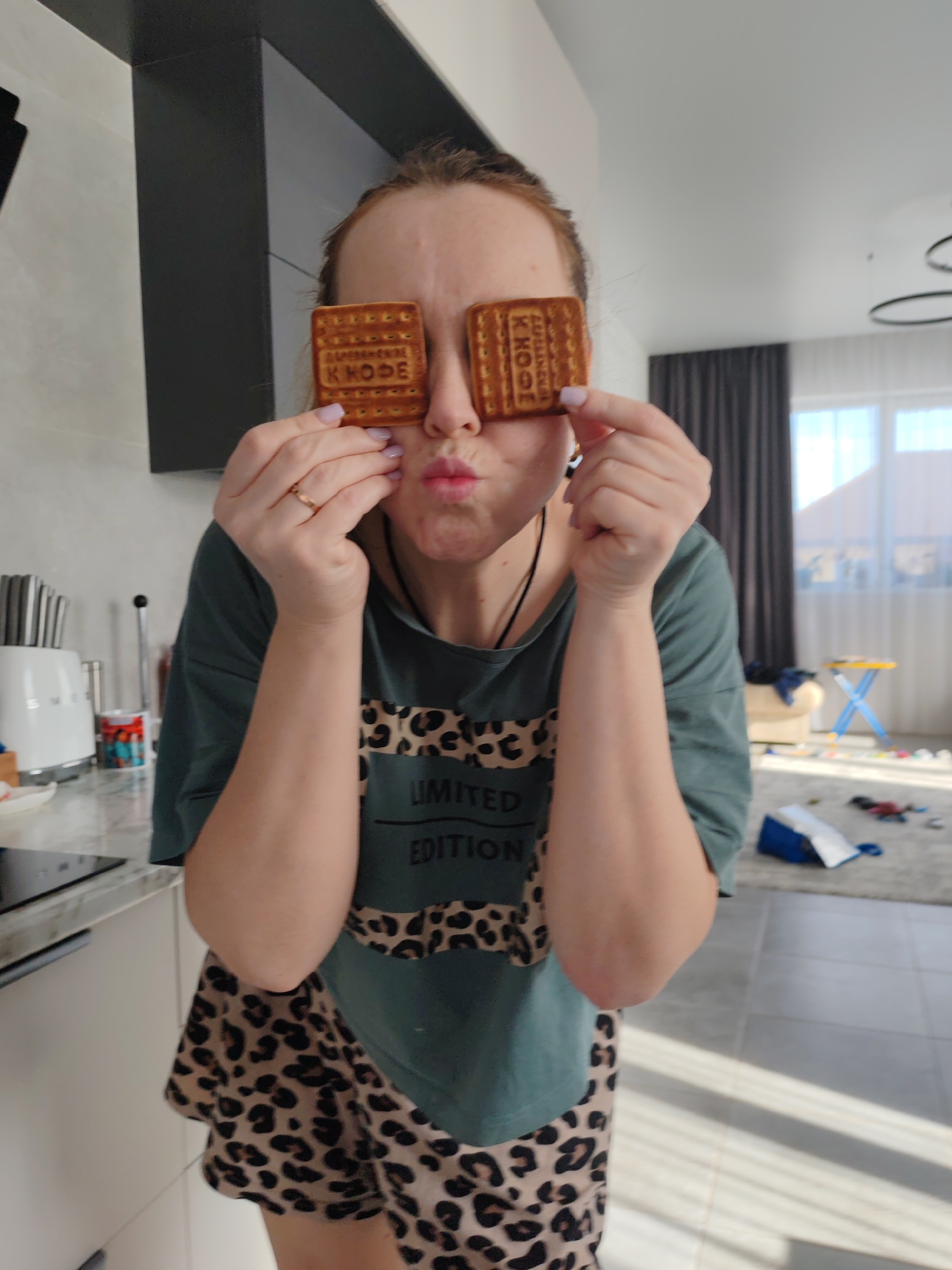 A female individual is depicted indoors, in what appears to be a residential kitchen and living area in the city of Chaikovsky, Russia. She stands facing the camera, holding a rectangular biscuit over each eye with her hands, completely obscuring her vision. Her lips are puckered forward. She wears a dark teal t-shirt with a leopard print design and the text "LIMITED EDITION" across the chest, paired with leopard print shorts. A thin metallic ring is visible on her left ring finger, and her fingernails are painted a light purple-pink hue. The biscuits are light brown and rectangular, with Cyrillic text partially visible on their surface, including "КАЗАНСКИЕ ХЛЕБОБУЛОЧНЫЕ" and "K HODE".
To the left of the subject, a kitchen counter with a light gray marbled surface is visible, featuring a white appliance housing several knives, a red and white striped mug, and other kitchen items. White cabinetry is positioned below the counter. To the right and behind the subject, a living area with light gray tiled flooring is illuminated by natural light entering through a large window. Dark gray drapes frame the window. Within this area, a light-colored armchair, a small yellow-topped table with blue legs, and various scattered items including a blue bag and colorful objects are visible on the floor. A modern, dark-colored circular light fixture is partially visible on the ceiling.
