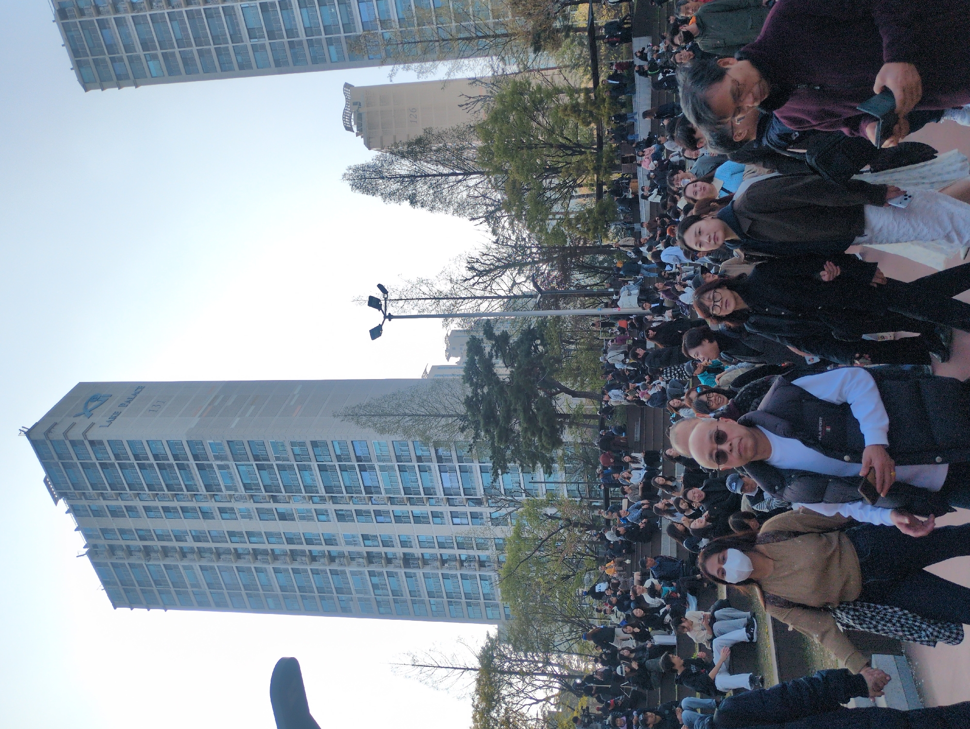 A large crowd of people, mostly Asian in appearance, stand and sit in an outdoor plaza, near the Lotte Palace apartment building located at 137 Seosomun-ro in Seoul, South Korea. Many are holding phones. A street lamp is visible among sparse trees. The overall view is tilted to the left.