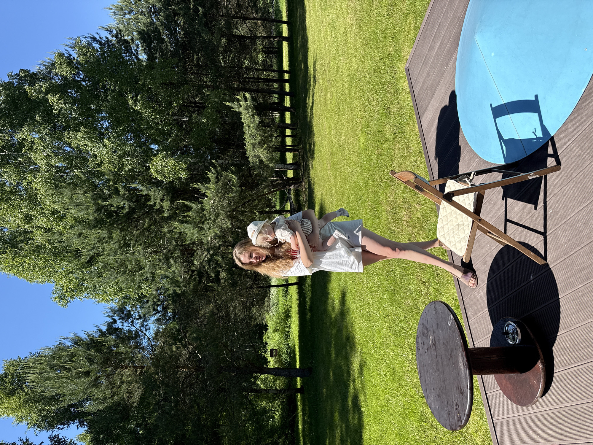 The image captures an outdoor scene in Копнино, Russia, on a bright, sunny day. An adult female, with long blonde hair and wearing a white t-shirt and shorts, stands on a vibrant green lawn. She is holding a baby, who is dressed in a light-colored bucket hat, a striped top, and shorts. Both individuals are looking towards the right side of the frame. In the immediate foreground, a dark brown wooden deck is visible, featuring a round wooden table with an ashtray on its lower tier, a wooden folding chair with a light-colored cushion, and a large, partially visible blue oval object. The background consists of a dense treeline composed of tall pine trees and deciduous trees with light bark, indicative of birches, bordering the expansive green lawn. A clear blue sky is visible above the trees, and strong sunlight casts pronounced shadows from objects on the deck.