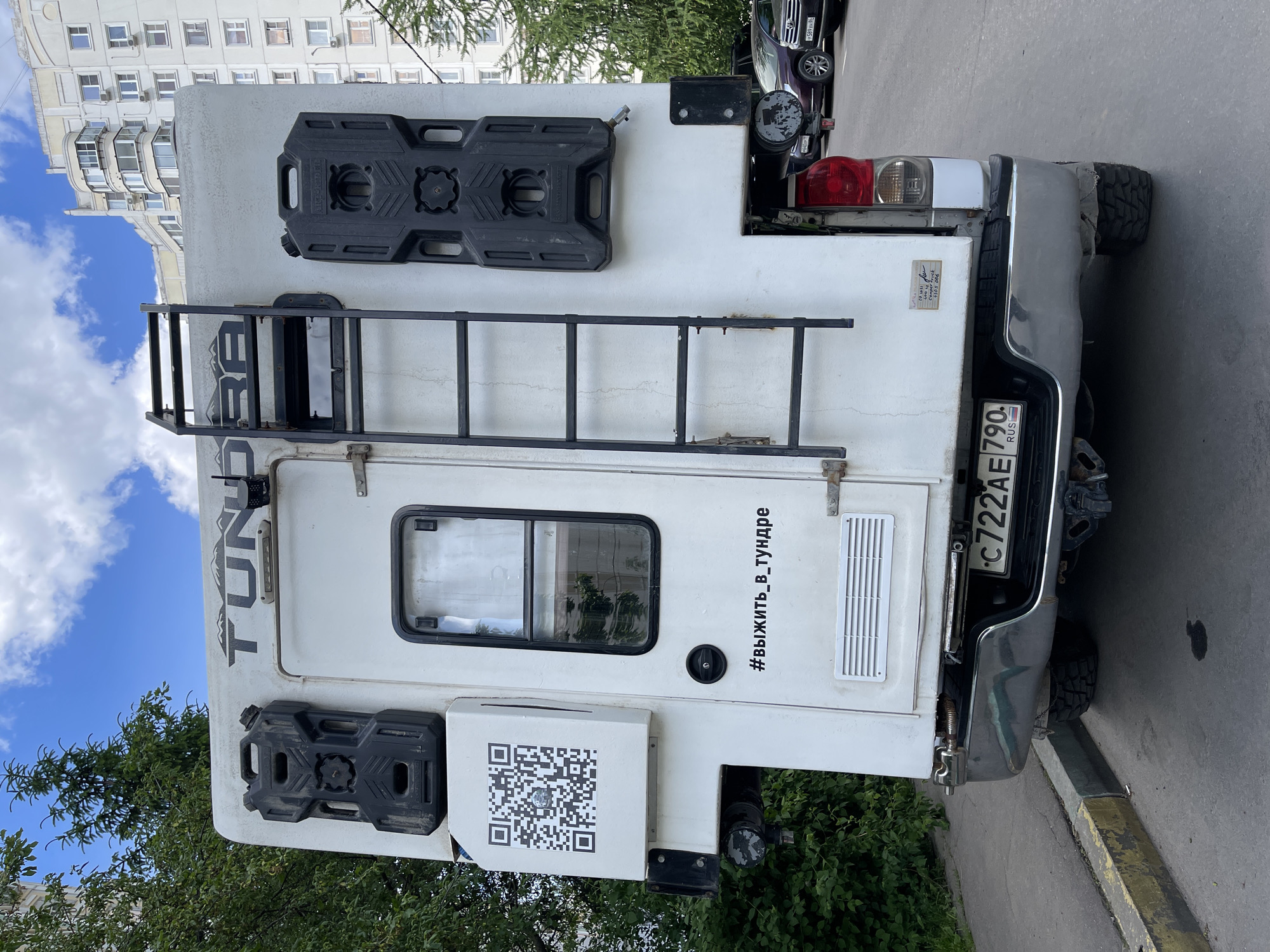 A white camper truck with Russian license plate C722AE790 is parked on a street next to a curb. The truck is equipped with a ladder, two black gas canisters, and a QR code sticker. The phrase "#выжить_в_тундре" is written on the side, translating to "#survive_in_the_tundra." In the background, an apartment building is visible, suggesting the scene is located in a city, identified as Moscow, Russia.
