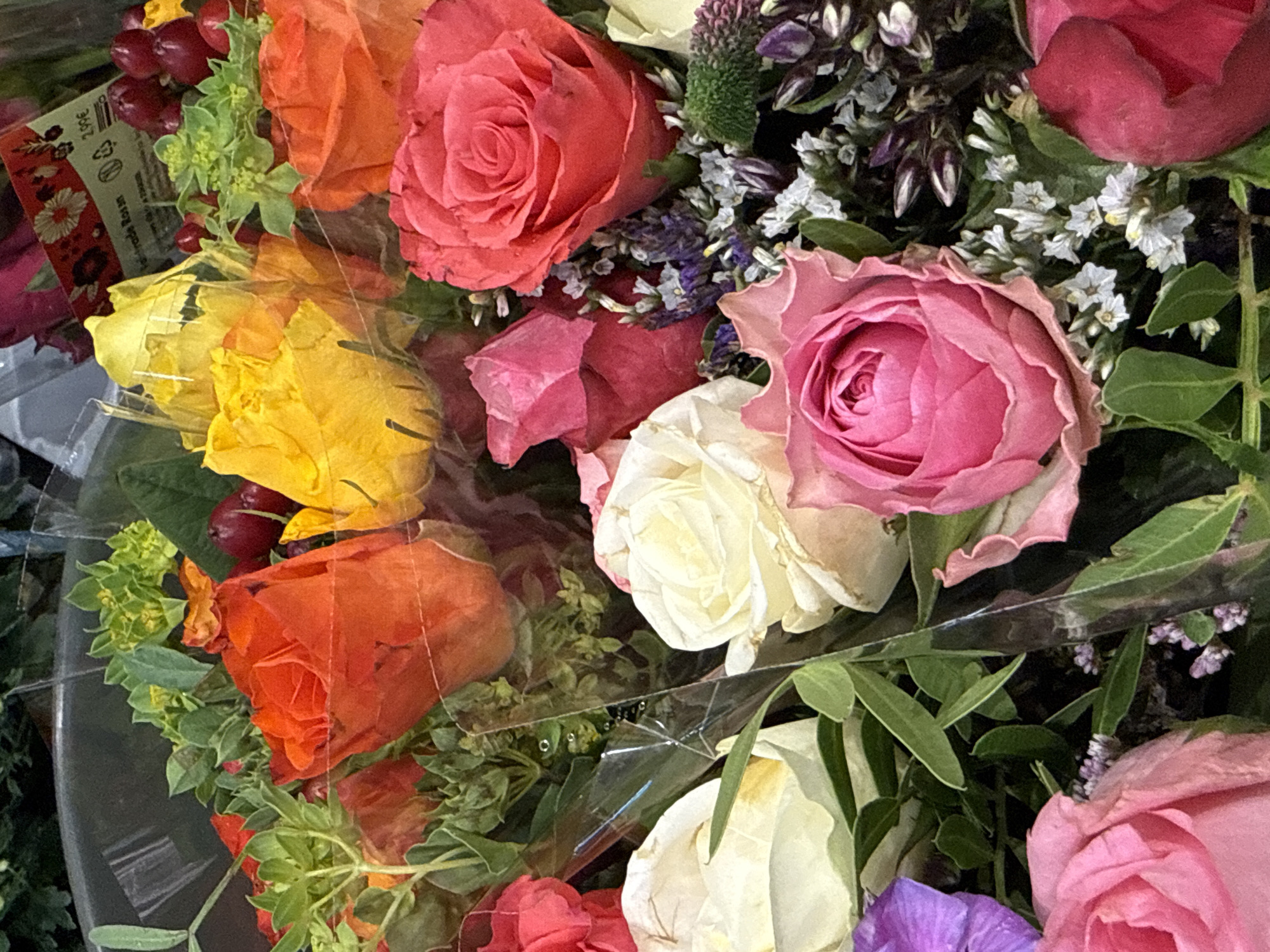 This is a close-up, eye-level, indoor photograph of a bouquet of roses and other flowers. The bouquet is predominantly composed of roses in various colors, including vibrant orange, bright yellow, deep pink, light pink, and white. Interspersed with the roses are small clusters of green foliage and delicate, light purple and white filler flowers. Some dark red berries are also visible. The flowers are arranged in a dense cluster and appear to be wrapped in clear plastic. A small white label with German text and a price of "2.99€" is partially visible at the top right of the image, suggesting the bouquet is for sale. The background is dark and out of focus, providing no specific environmental context beyond the implied setting of a flower shop or market. No people are present in the frame. The image captures the textures and colors of the floral arrangement. The location of Bad Lauterberg im Harz, Germany, is not discernible from the visual elements of the photograph itself.