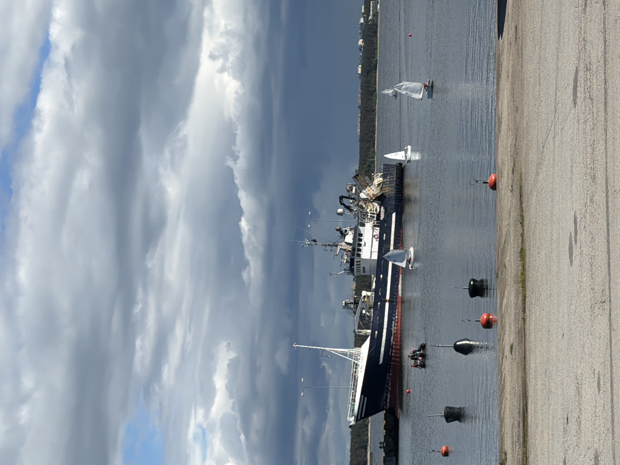A large dark blue and white fishing trawler is docked at a pier in the foreground, with a dense line of trees and buildings visible in the background. Several small sailboats are scattered across the water, some with sails up, suggesting a leisurely activity. A group of people are in a small inflatable boat near the trawler. The sky is partially cloudy, with dramatic dark clouds dominating the upper portion of the frame, contrasting with patches of lighter sky and the sun illuminating parts of the water and foreground. Buoys are visible in the water between the shore and the boats. The location is the harbor of Viimsi, Estonia.