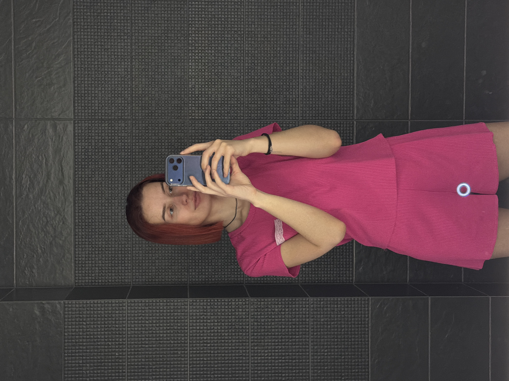 A person is taking a selfie in what appears to be a bathroom. The person is wearing a pink outfit and holding a blue smartphone. The bathroom has dark gray tiles on the wall. The exact location in Chaikovsky, Russia cannot be determined from the image.