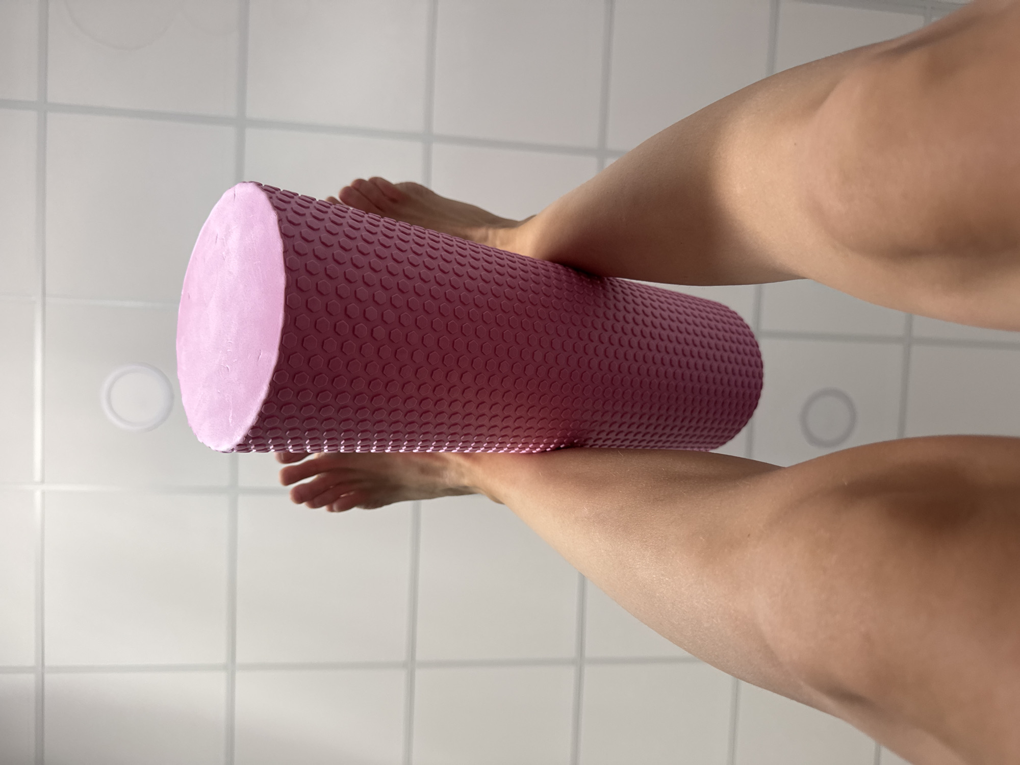 The image shows a person's legs and feet positioned above a pink, textured foam roller. The roller is cylindrical and has a honeycomb-like pattern on its surface. The person's feet are bare and rest on the top of the roller, with their toes visible. The legs are bent and positioned to the sides of the roller. The background consists of white tiled walls, likely in a bathroom or a similar tiled room. The lighting suggests it is daytime. There is a circular light fixture visible on the wall in the background. The overall impression is that the person is using the foam roller for a massage or stretching exercise.