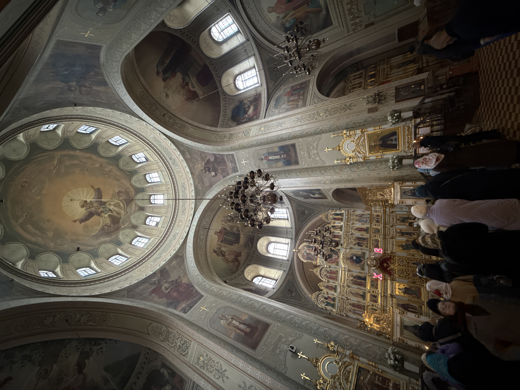 Inside the Church of St. Sergius of Radonezh, Sviyazhsk, Russia, a high-angle shot reveals the ornate interior with religious frescoes and paintings adorning the walls and ceilings. Chandeliers hang from the ceiling. People are gathered, some kneeling in prayer before a golden iconostasis. The architecture showcases arches, windows, and detailed designs, characteristic of Orthodox churches.