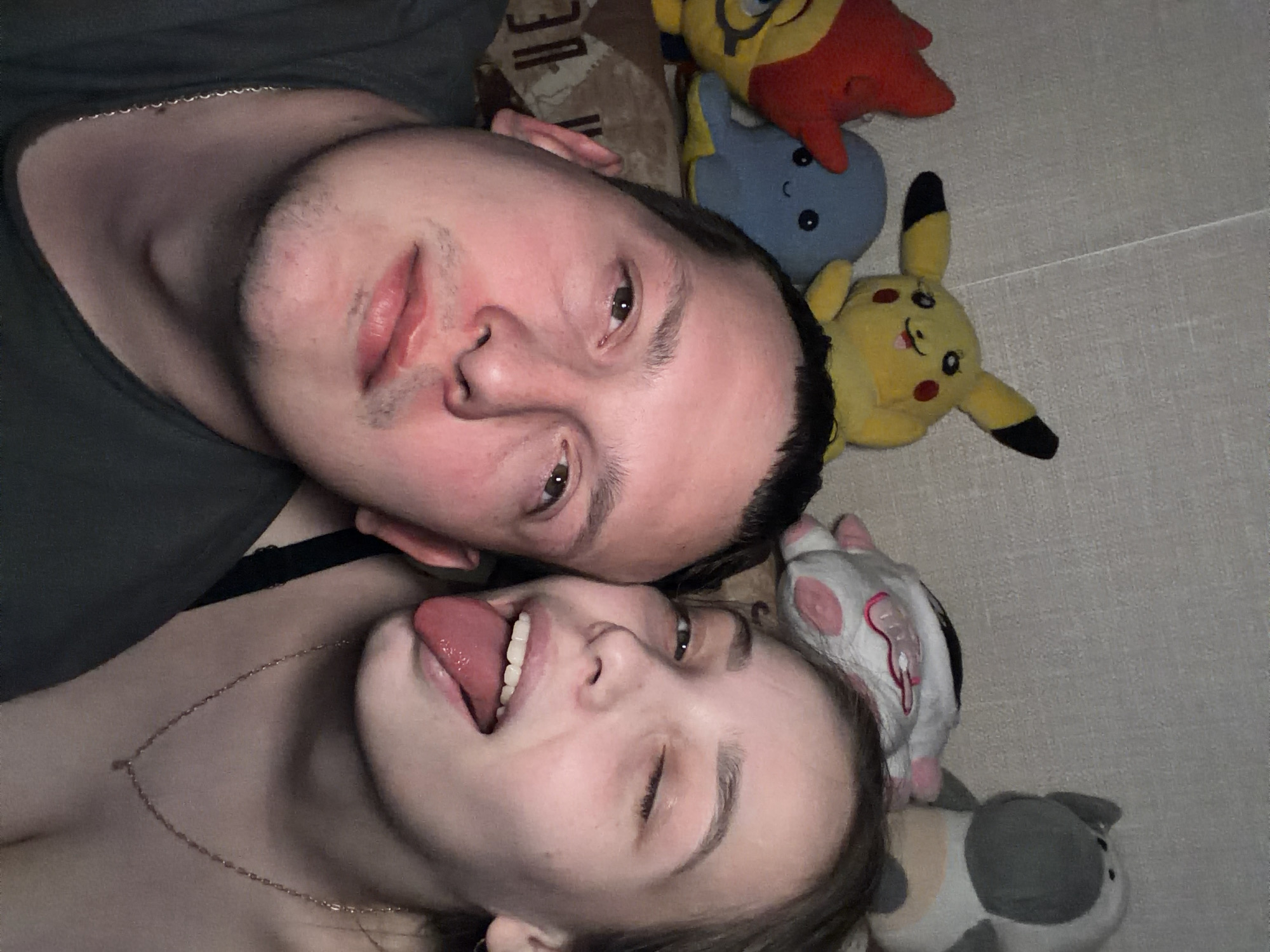 This media file captures a close-up, top-down view of two individuals, a man and a woman, lying close together indoors. The photo appears to be a casual selfie, taken in Chaikovsky, Russia.
The man is positioned at the top left, wearing a dark t-shirt, with short dark hair and light stubble on his chin and upper lip. He looks directly at the camera with open eyes and a neutral expression. Adjacent to him, the woman lies with her head near his shoulder. She has long, light brown hair, and is wearing a dark top, possibly revealing a bra strap. A delicate gold chain necklace rests on her collarbone. She looks playfully towards the camera, with one eye slightly squinted or winking, and her tongue sticking out.
Around and behind them, several plush toys are scattered on a light-colored surface, possibly a bedspread or carpet. Visible toys include a yellow Minion, a red and orange creature (resembling Charmander), a blue creature, a yellow Pikachu, a white and pink cat-like plush, and a grey and white spotted cat-like plush. The background under the man's head shows a darker, patterned fabric.
The overall scene conveys a relaxed and playful, intimate moment between the two individuals, likely friends or a couple. The indoor setting suggests a comfortable, personal space. There is no visible text in the image.
