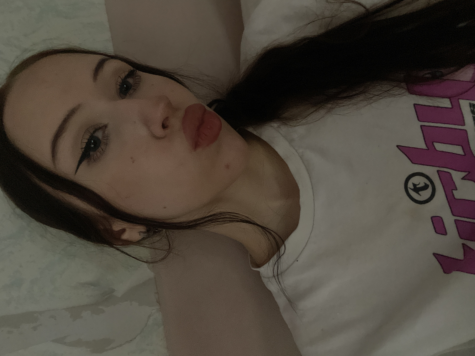 The image is a close-up selfie of a young woman with dark hair, wearing a white t-shirt with pink lettering. She is looking at the camera with her lips puckered. She has winged eyeliner and her hair is pulled back in a ponytail. The background appears to be a wall with a patterned wallpaper. There is no discernible information about the city of Votkinsk, Russia in this image.