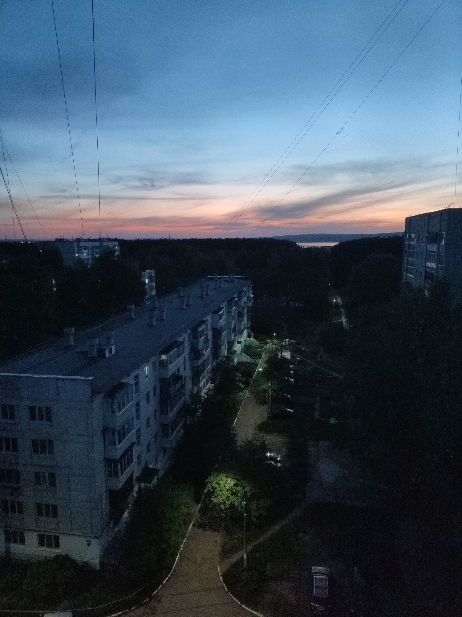 Here is a description of the image:
The image shows a nighttime urban scene. In the foreground, there are large, gray apartment buildings of Soviet design. The buildings have rows of windows and balconies. A paved path leads away from the building in the lower left corner, illuminated by a street light. Several parked cars are visible along the path. In the distance, there is a tree line and a body of water, possibly a lake or river. The sky is a gradient of blue and orange, indicating sunset or sunrise. Power lines are strung across the sky. There are no people visible in the image. The overall scene is quiet and somewhat dark.