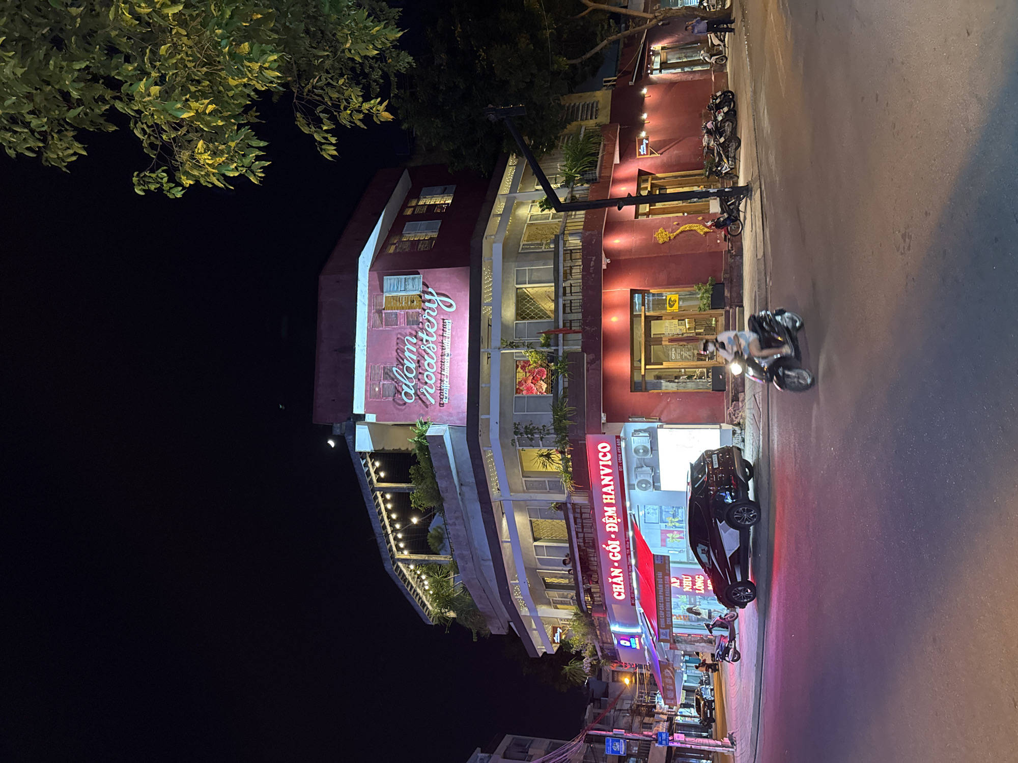 The image depicts a street scene in Nha Trang, Vietnam at night. A multi-story building with a prominent red facade and a sign reading "clam roastery" is the central focus. The building features several levels with balconies and outdoor seating areas illuminated by string lights. Various shops are visible on the ground floor, including one with a red awning and the sign "CHAN - GOI - DEM HANVICO." A black SUV is parked on the sidewalk in front of one of the businesses. Several motorbikes are parked along the street and sidewalk. A few people are visible; one individual is riding a motorbike in the foreground, another appears to be walking near a shop entrance, and some individuals are seated at outdoor tables on the building's balconies. The street is paved and appears wet in some areas, reflecting the lights. Trees and streetlights are also present in the scene. The sky is dark, indicating nighttime, with a faint crescent moon visible.