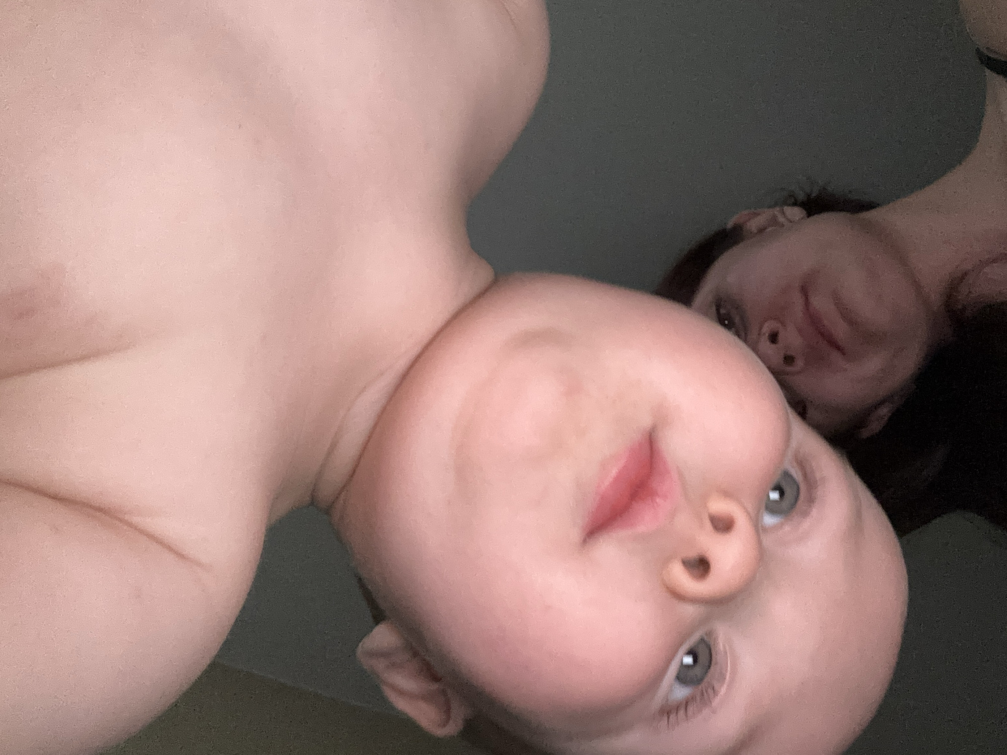 The image shows an upside-down, close-up view of two individuals. In the foreground, a baby's face is prominently featured, with its eyes, nose, and mouth visible. The baby has fair skin and light-colored eyes. Behind the baby, partially obscured, is the face of an adult, likely female, with darker hair and visible skin on her forehead and cheeks. The lighting in the image is dim, creating a shadowy effect. The background appears to be a plain, dark surface, possibly a wall or a piece of furniture. The orientation of the image suggests it was taken from above, looking down at the subjects. There is no discernible information within the image to indicate its specific location in Naberezhnye Chelny, Russia.