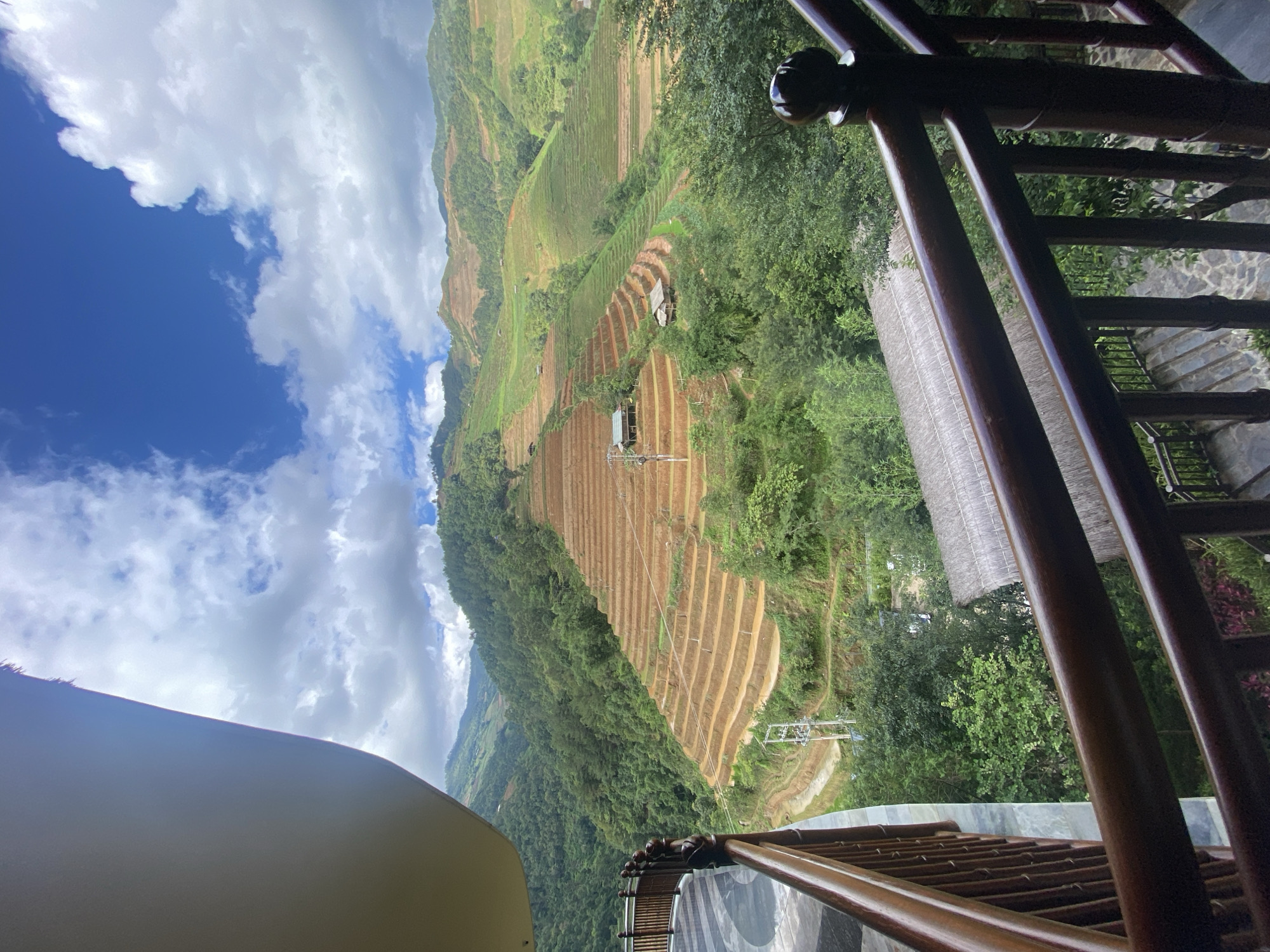 This image captures a panoramic view of a mountainous landscape in Púng Luông, Vietnam, observed from an elevated architectural structure, likely a balcony or terrace. The sky is partially blue with prominent white and grey cumulus clouds.
In the mid-ground and background, multiple mountain ranges are visible, densely covered in verdant trees and shrubs. Extensive agricultural terracing dominates the slopes; some terraces appear freshly tilled, revealing brown earth, while others are covered in green vegetation, indicative of crops. Several small, traditional-style huts or houses with thatched roofs are sparsely distributed across these terraced fields. A utility pole with power lines is discernible within the terraced area on the left side of the mid-ground.
In the immediate foreground, dark brown wooden railings, consistent with a balcony or staircase, frame the lower and right portions of the image. The railing on the left is curved, and a grey and black patterned tiled floor is visible behind it. The upper left corner shows a curved, light-colored ceiling or overhang of the viewing structure. Below the right-hand railing, steps and a stone-paved pathway can be seen descending into dense foliage, with a small section of a thatched roof visible through the trees. No individuals are present in the scene.