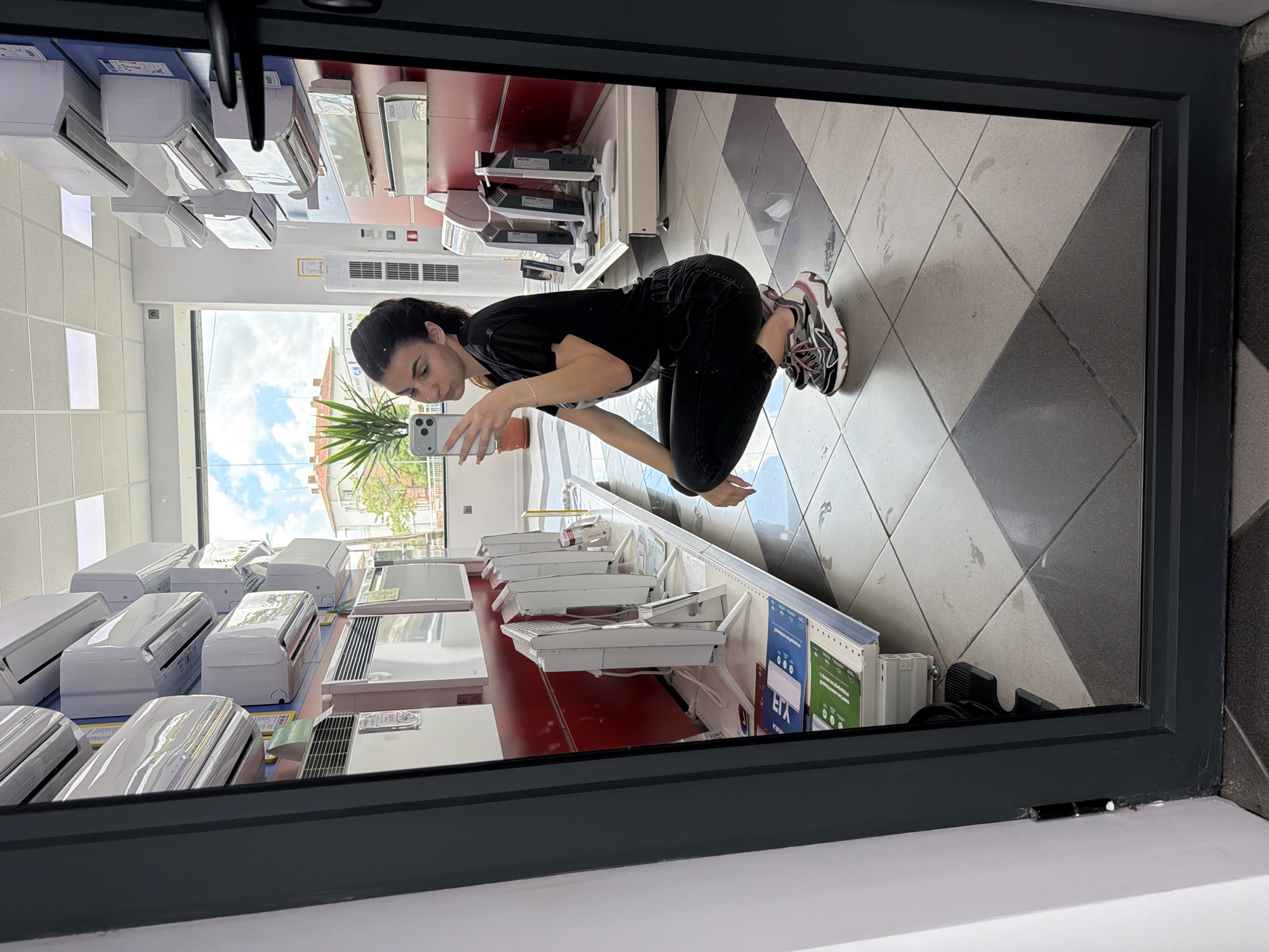 A woman is visible in a mirror reflection, kneeling in what appears to be a store or showroom environment. She is holding a phone and seems to be taking a picture. The store is filled with air conditioning units, suggesting an appliance or electronics store. Outside the window, there's a view of a street scene with greenery and buildings, suggesting an urban or suburban setting. The store has a tiled floor and appears to be well-lit. Based on available visual clues, I can’t confirm the location in Gabrovo, Bulgaria.