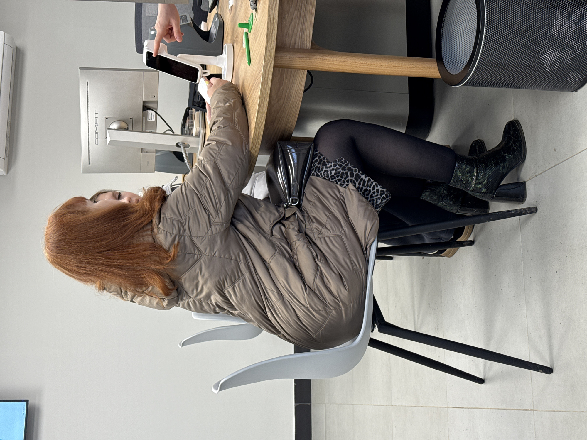 A woman with long red hair wearing a beige quilted jacket, black tights, a leopard print skirt, and green patterned boots is sitting in a white plastic chair at a light wood table. She is interacting with a person whose hand is visible and holding a white mobile device. A desktop computer monitor with the word "COMPIT" is visible behind them. A cylindrical air purifier is on the right side of the frame. The setting appears to be indoors, possibly a retail or office environment, though the exact location in Votkinsk, Russia, is not discernible from the image.