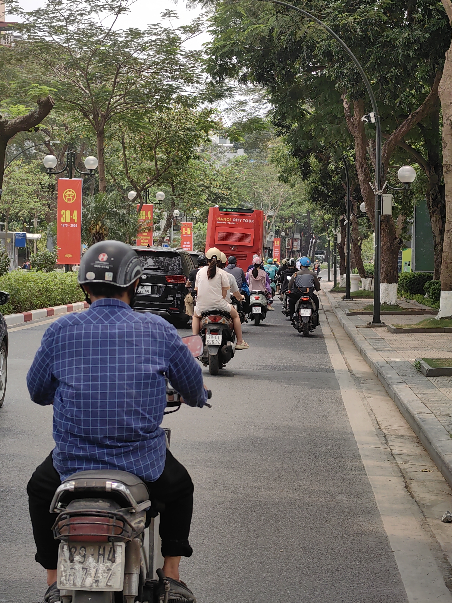 A medium, eye-level shot depicts a street in Hà Nội, Vietnam, filled with a line of motorbikes and scooters moving in the same direction. The viewpoint is from behind a motorbike rider, wearing a blue checkered shirt and a helmet. The motorbike's license plate reads "29-H4 16742".
Ahead, a dark KIA SUV is followed by multiple motorbikes carrying riders and passengers. A red "Hanoi City Tour" bus is further up the street. Red banners with the text "30-4 1975-2026" are posted along the street. The license plate of the vehicle ahead reads "30H 537.95" and another license plate of a vehicle reads "29-C1 696.34".
The street is lined with trees and decorative lampposts. A sidewalk with light-colored pavement runs along the right side of the street. The weather appears overcast.