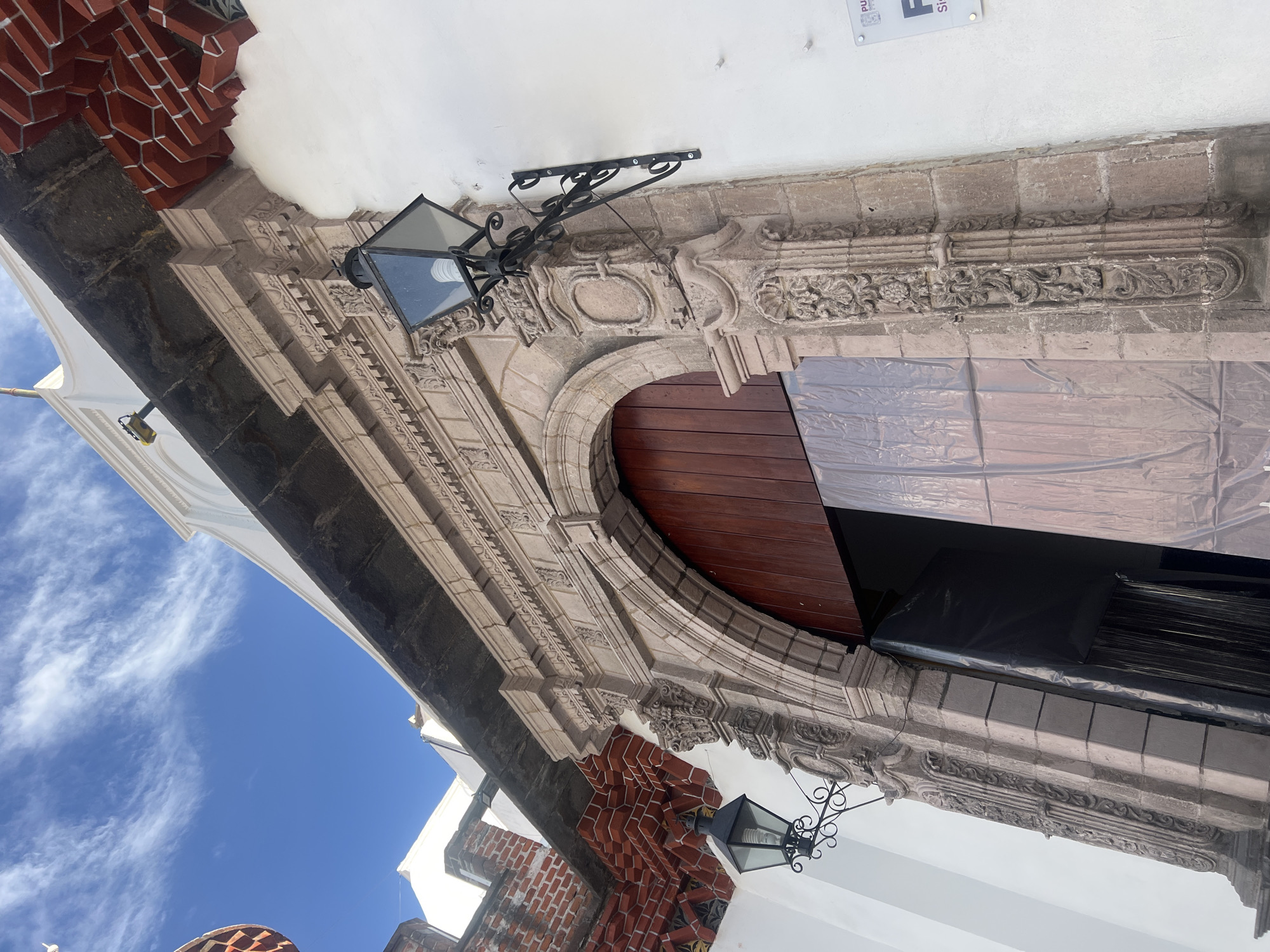 A ground-level shot shows an ornate stone entryway in Puebla, Mexico. The light-stone archway is flanked by red brick on the left and right. A black metal lamp fixture hangs from the stone archway above the brown wooden door. An ornate carved stone border stretches across the top of the entryway. The sky, which is visible from the left side of the image, is blue with white clouds.