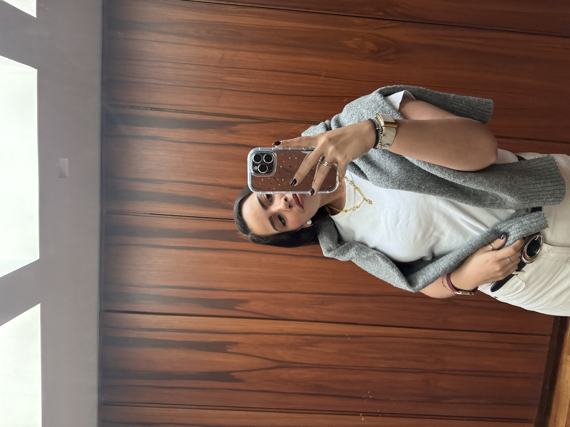 A woman stands indoors against a wooden wall, taking a selfie with her smartphone. The phone has a transparent case adorned with small, colorful dots. She is partially obscuring her face with the phone. She is wearing a white t-shirt, a gray cardigan draped over her shoulders, and white pants. Jewelry includes a gold watch, bracelets, rings, and a delicate gold necklace. The environment includes a window on the left side, allowing natural light into the space. It is impossible to verify the location is San Bernardino Tlaxcalancingo, Mexico, given the lack of contextual details.