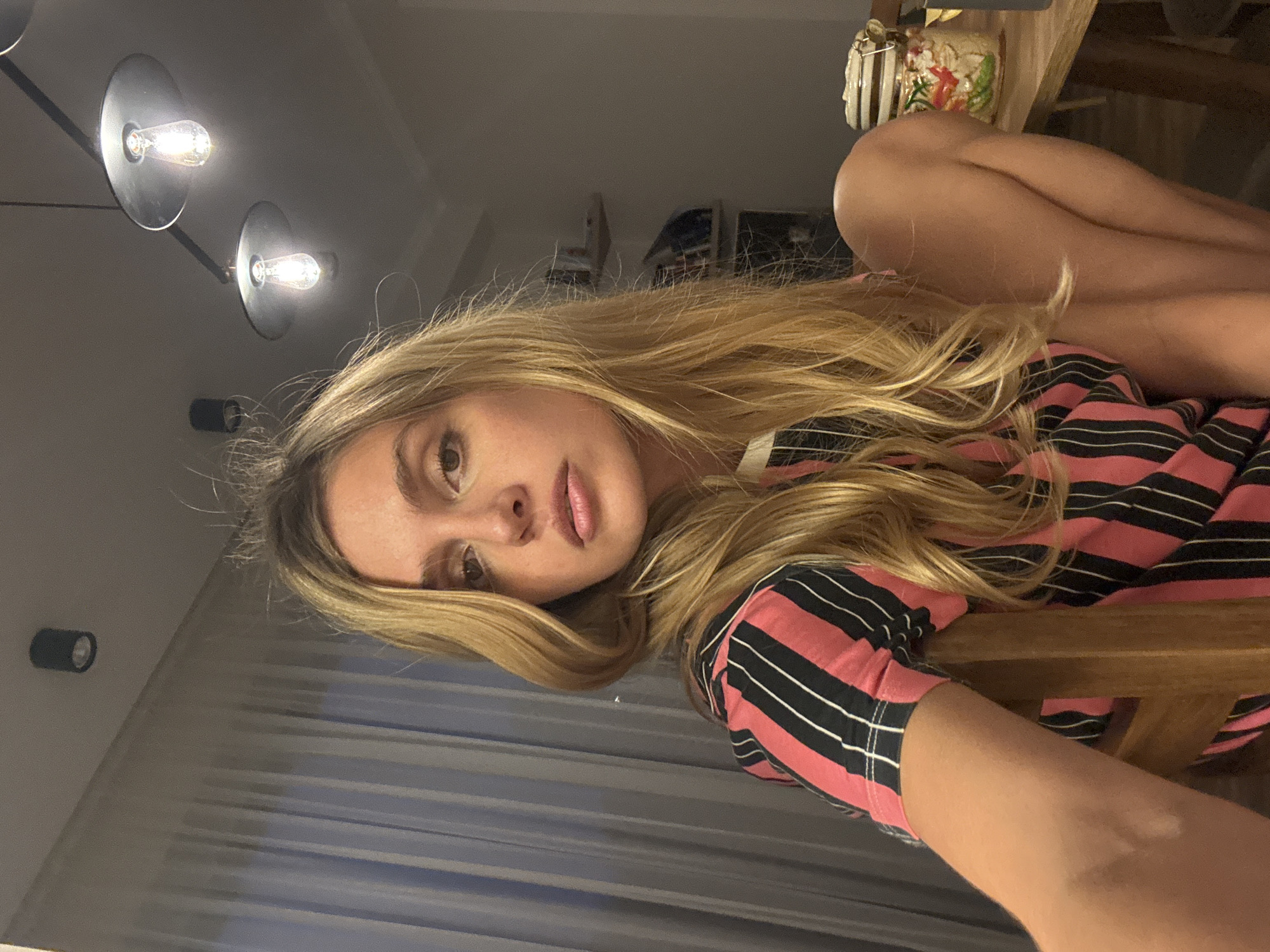 The image, rotated 90 degrees to the left, captures a young woman with long, wavy blonde hair and fair skin looking directly at the camera. Her expression is neutral, with slightly parted lips. She is wearing a short-sleeved top with horizontal stripes of pink and black, accented by thin white lines. Her arm and parts of her legs are visible, suggesting she is reclining or leaning back, with a wooden chair's back seen behind and next to her.
The setting is indoors, likely a home, during the evening or night, illuminated by artificial lighting. In the background, a modern ceiling light fixture with two glowing Edison-style bulbs casts a warm light. A smaller, black cylindrical light fixture is also visible on a wall further back. To the upper right, on what appears to be a table or shelf, there are decorative objects, including a container holding green foliage. No text is visible in the image.