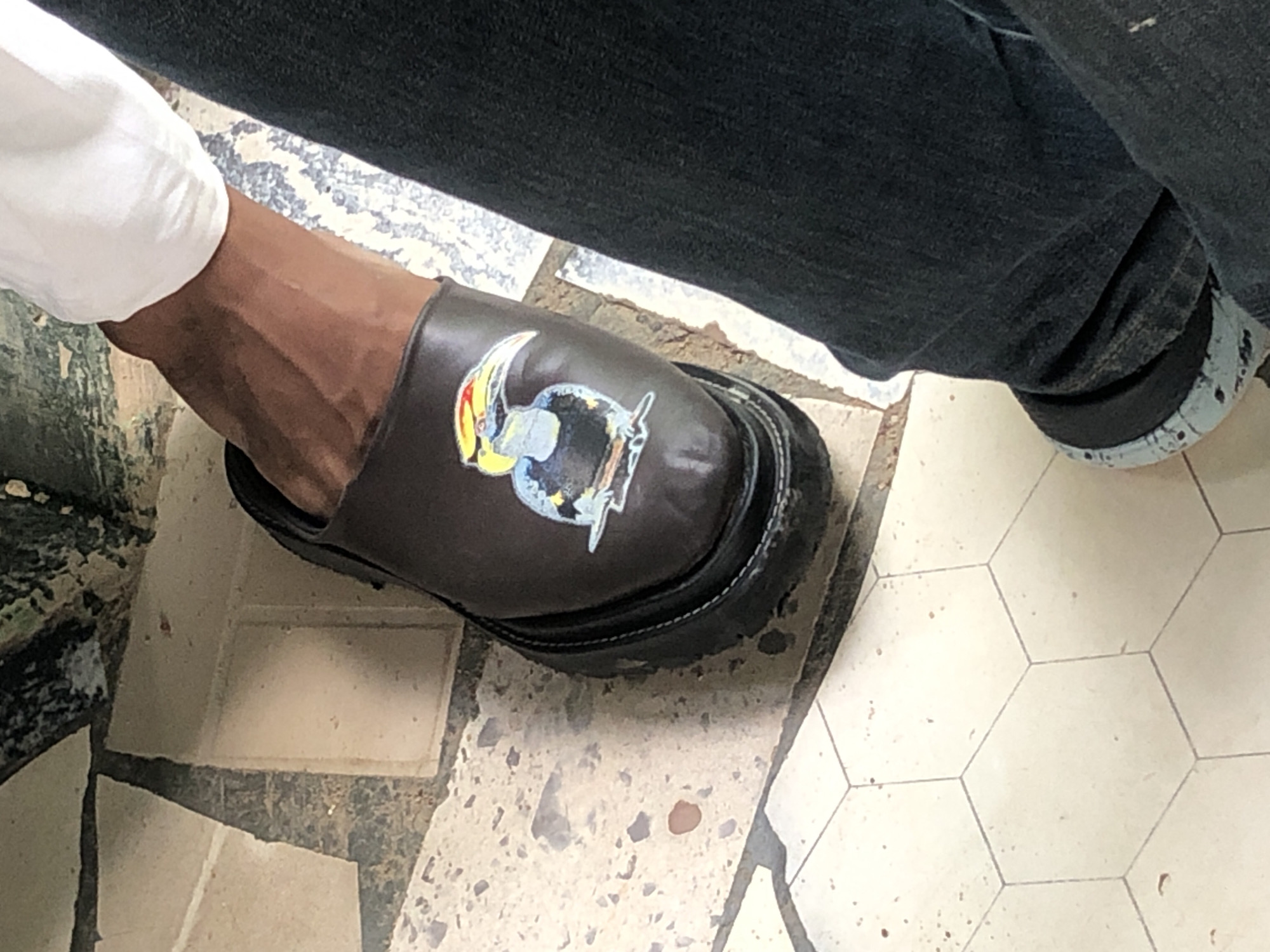 The image shows a close-up, top-down view of a person's left foot wearing a brown slip-on shoe decorated with a colorful sticker of a hornbill bird. The shoe has a thick, black platform sole. The person is wearing dark denim jeans. The background consists of two different types of tiled flooring. One is a light-colored hexagonal tile, and the other appears to be larger, irregular stone or tile pieces with visible grout lines and some discoloration. The scene is well-lit, suggesting it was taken outdoors or in a brightly lit indoor space. The image quality is clear, allowing for detailed observation of the shoe, the sticker, and the flooring.