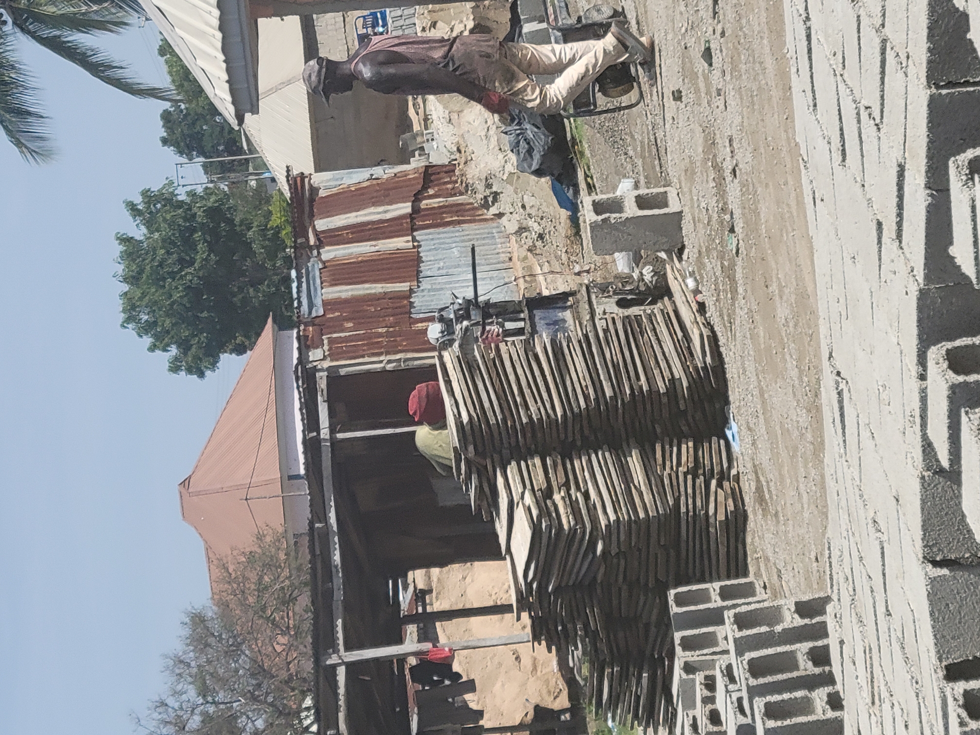 The image depicts an outdoor scene in Kwamba, Nigeria, likely a construction or material yard. In the foreground, stacks of grey concrete blocks are visible, suggesting ongoing construction. A large pile of flat, wooden planks is stacked to the left of the frame, next to a rustic, open-sided shelter with corrugated metal roofing and some clothing hanging from its structure.
A man, dressed in a light-colored sleeveless shirt and pants, wearing a cap, and with a bandana or cloth tied around his forearm, is walking from left to right in the mid-ground. He appears to be wearing gloves and is moving past a generator. Behind him, there is more corrugated metal sheeting forming part of a structure, and beyond that, trees and buildings under a clear, bright sky. The ground is a mix of dirt and gravel, typical of an undeveloped or industrial area. The overall impression is one of a busy, functional outdoor workspace.