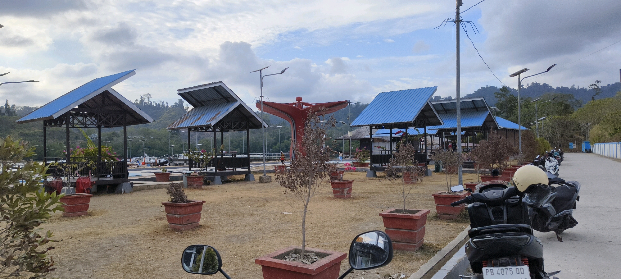 The image depicts a waterfront area in Waisai, Indonesia. In the foreground, two parked motorcycles are visible, one with a light-colored helmet on its seat. The ground is covered with dry, light-brown earth and scattered with several large, red square planters, some containing small trees or bushes.
A paved walkway runs along the right side of the image, lined with more parked motorcycles. Beyond the paved area, a series of open-sided gazebos with blue corrugated metal roofs are arranged. These gazebos appear to be part of a public space or resting area, with some featuring potted plants.
In the mid-ground, a distinctive red abstract sculpture stands prominently, resembling a stylized bird or a flower. A pedestrian walkway or pier extends into the water behind the gazebos. Several boats are docked in the water.
The background is dominated by lush green hills and mountains covered with dense tropical vegetation. The sky is overcast with large, white clouds. The overall impression is of a semi-developed coastal public area with recreational facilities, set against a natural, hilly landscape.
