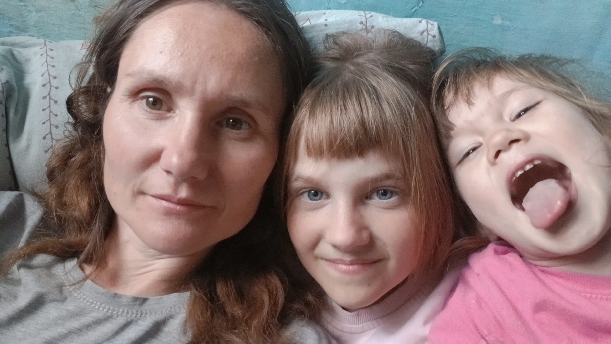 A close-up selfie shows three individuals, a woman and two young girls. The woman, on the left, has brown hair and light-colored eyes. She is wearing a gray t-shirt. To her right is a girl with blonde hair and blue eyes. She is smiling slightly. On the right edge of the frame, a second girl with brown hair and a pink shirt is sticking her tongue out and has her mouth open. The subjects appear to be lying down. The environment includes a pillow with a light-colored pattern. The location is Unknown, Unknown.