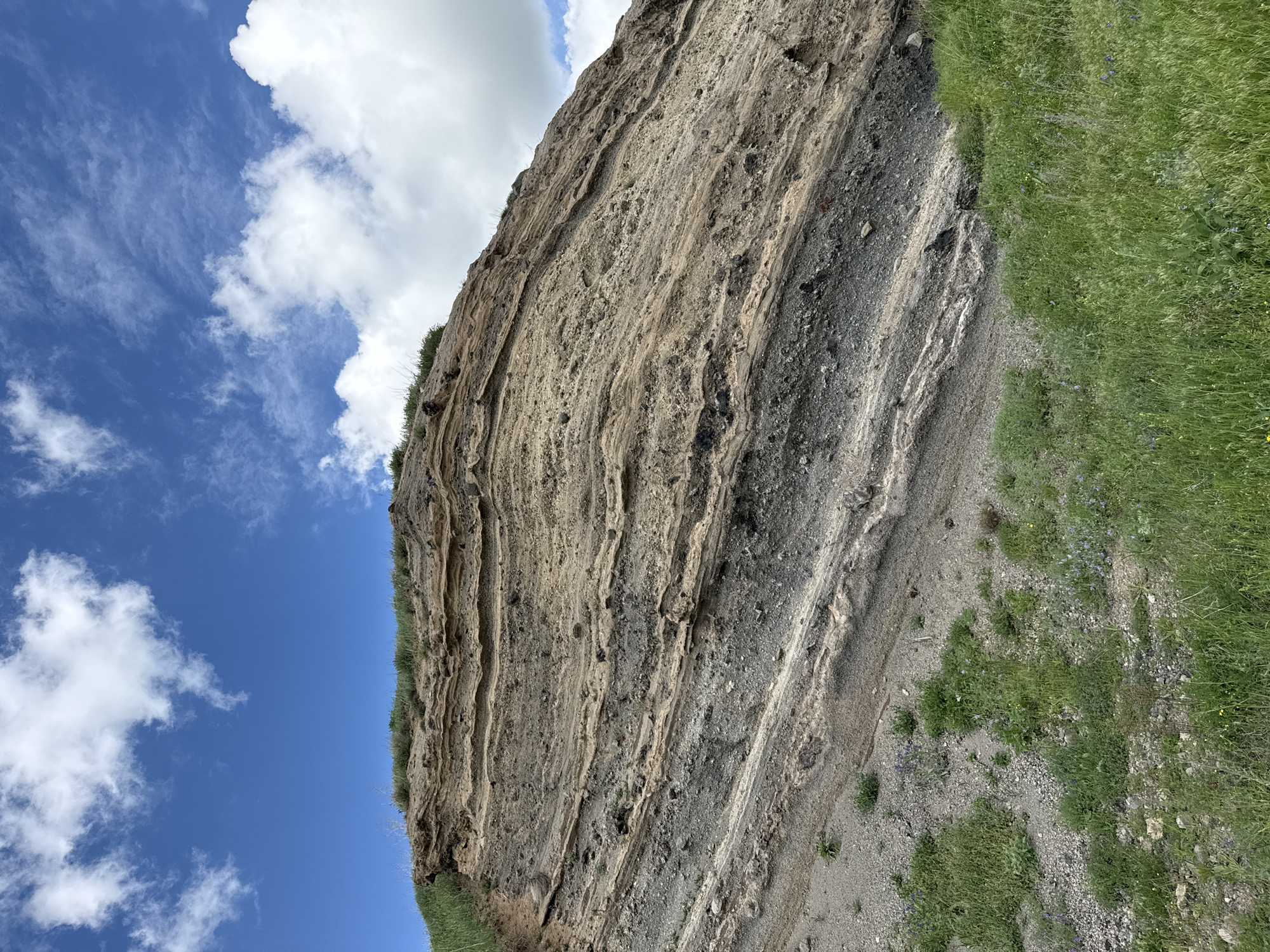 The image shows a geological formation composed of layered volcanic rock. The layers vary in color from light brown to dark gray and appear to be tilted. The formation is partially covered by green grass and some small plants, suggesting an outdoor, natural environment. The sky above is blue with scattered white clouds. The scene appears to be a natural landscape rather than an urban setting.