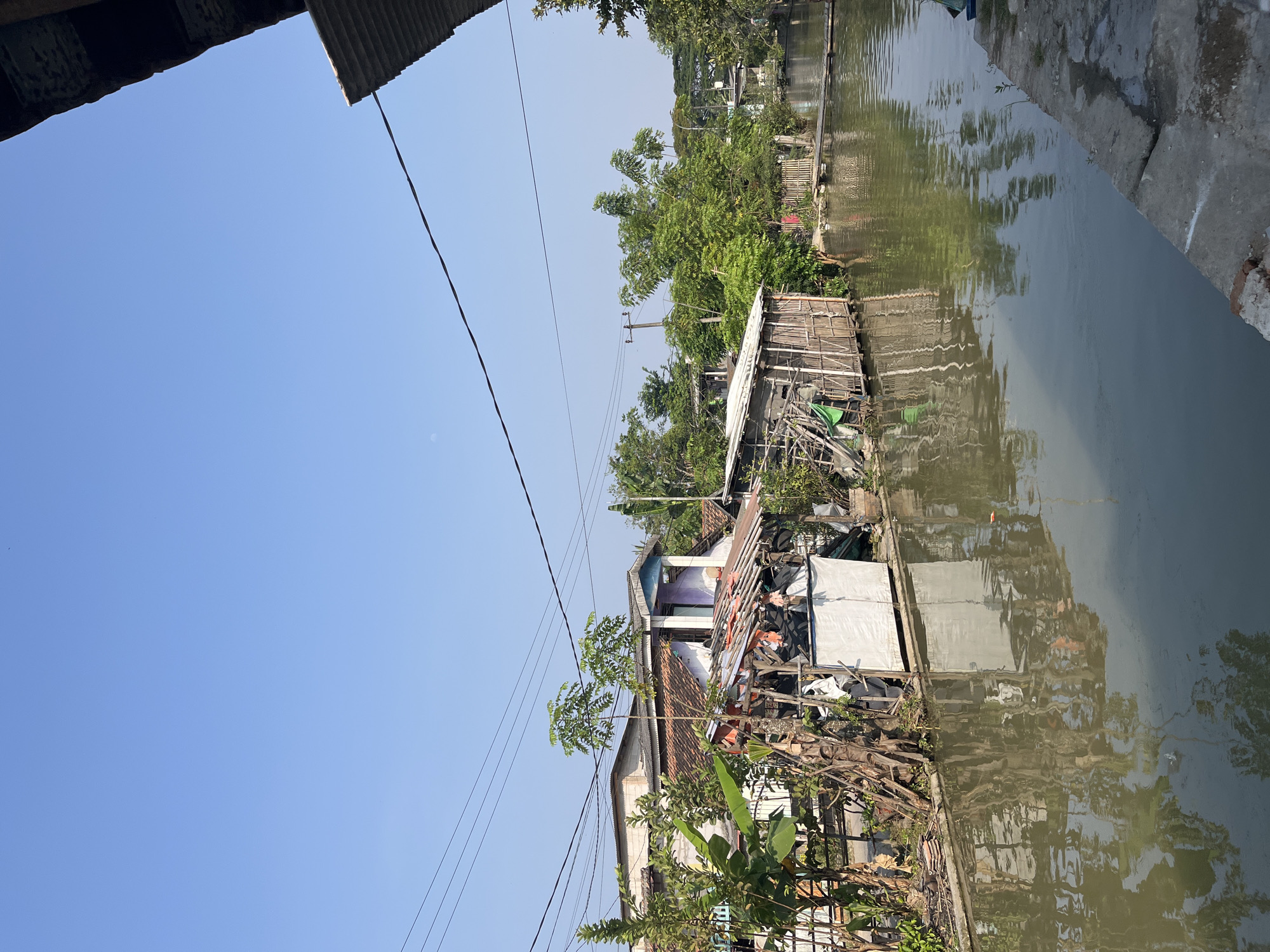 This image depicts a scene in Laban, Indonesia, showing a body of water, likely a canal or river, flowing alongside a row of buildings and vegetation. The water is a murky brown color and reflects the structures and trees on its banks.
On the left side of the water, there are several buildings. The closest building appears to be a makeshift structure with a roof made of corrugated metal and bamboo walls. Next to it is a more permanent building with a tiled roof and white walls, some parts of which are under construction or in disrepair. There is a lot of vegetation, including banana trees and other leafy plants, surrounding these structures.
Further down the waterway, more buildings and trees are visible, suggesting a densely populated area. Power lines stretch across the sky, indicating the presence of electricity infrastructure. The sky is clear and blue, with a faint moon visible.
The overall impression is one of a lived-in, perhaps less affluent, area of the city, characterized by its proximity to water and the coexistence of natural and built environments. There are no people visible in the image.
