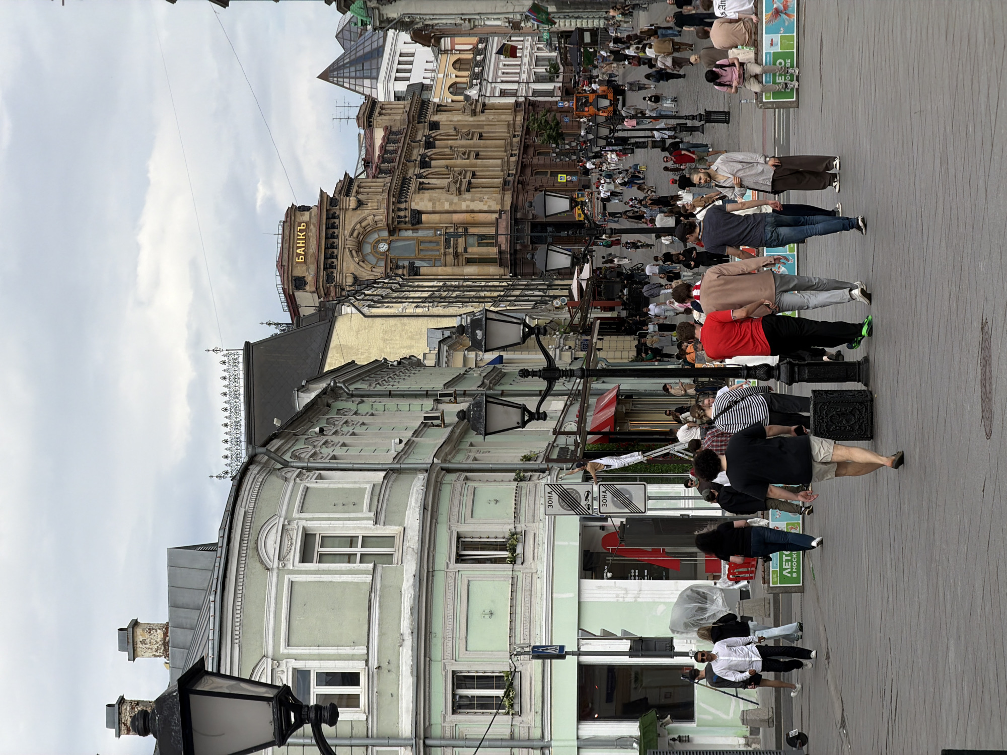 The image shows a street scene in Moscow, Russia. The street is crowded with pedestrians. Buildings line both sides of the street. One of the buildings has the word "БАНКЪ" (Bank) displayed in illuminated letters on its façade. Street lamps are visible along the pedestrian pathway. The sky is overcast with a light cloud cover.