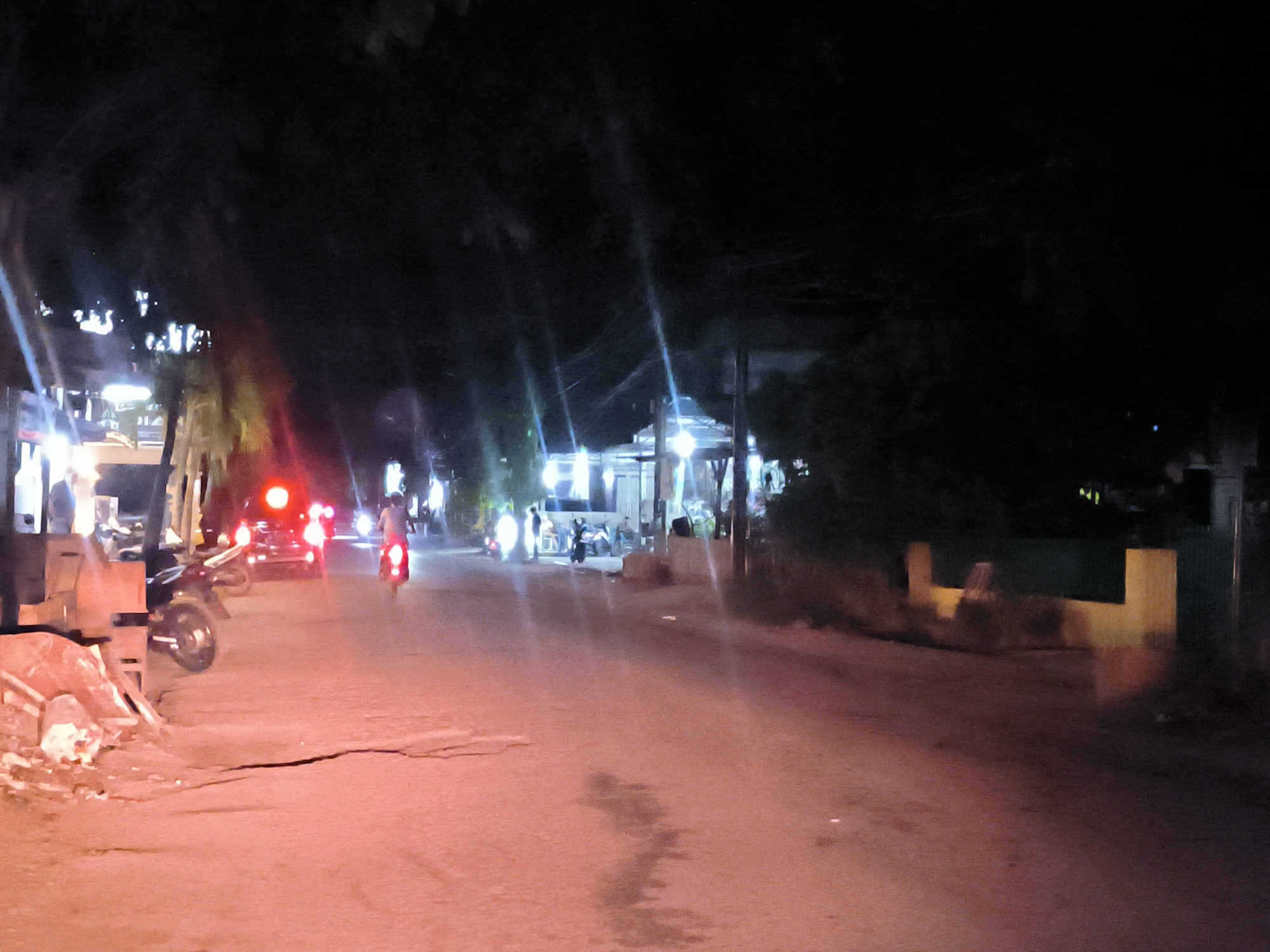 The image depicts a street at night in Langsa, Indonesia. The road is asphalt and appears to have some cracks and imperfections. On the left side of the road, there are construction materials, including stacked bricks and wooden planks. A parked motorcycle is nearby. Further down the street, two motorcycles are moving away from the viewer, with their red taillights visible. A building with bright lights is visible in the distance, and several people are gathered outside. The street is lined with trees and foliage on both sides, and the lighting is dim, with a reddish hue due to the artificial lights. The environment suggests a residential area.