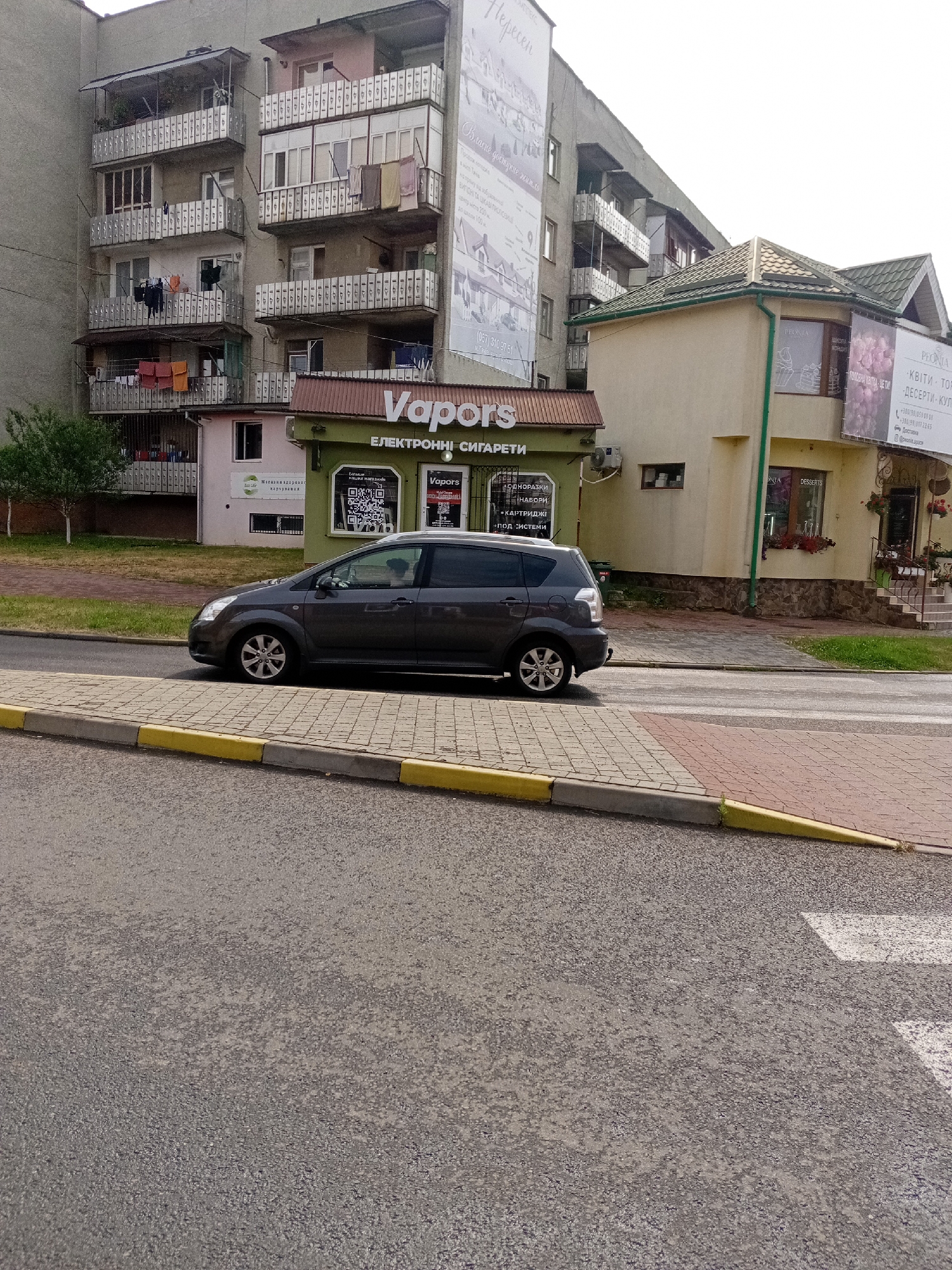 This is a street-level, eye-level, medium shot taken on a cloudy day, possibly in the late morning or early afternoon, in Tiachiv, Ukraine. The scene is dominated by buildings and a street with a car. On the left, a multi-story apartment building with many balconies, some with laundry hanging, takes up a significant portion of the frame. Below the apartment building, a small, light pink structure is partially visible.
To the right of the apartment building, a green storefront with the prominent text "Vapors" and "ЕЛЕКТРОННІ СИГАРЕТИ" (Electronic Cigarettes) is visible. This storefront has large windows displaying products and QR codes. Adjacent to the Vapors store is a beige building with a green roof, featuring a sign that reads "PEONIA" and lists services like "КВІТИ" (Flowers), "ТОР" (Cake), and "ДЕСЕРТИ" (Desserts). The phone numbers and social media handles are also visible on the PEONIA sign.
A dark gray car is parked on the street in front of the Vapors store, with its driver visible through the window. The street itself is paved, and a sidewalk made of interlocking bricks with yellow and gray curbs runs alongside it. The overall impression is of a typical urban street scene in a Ukrainian town, with commercial establishments lining the road. The weather appears overcast, suggesting a diffused light.