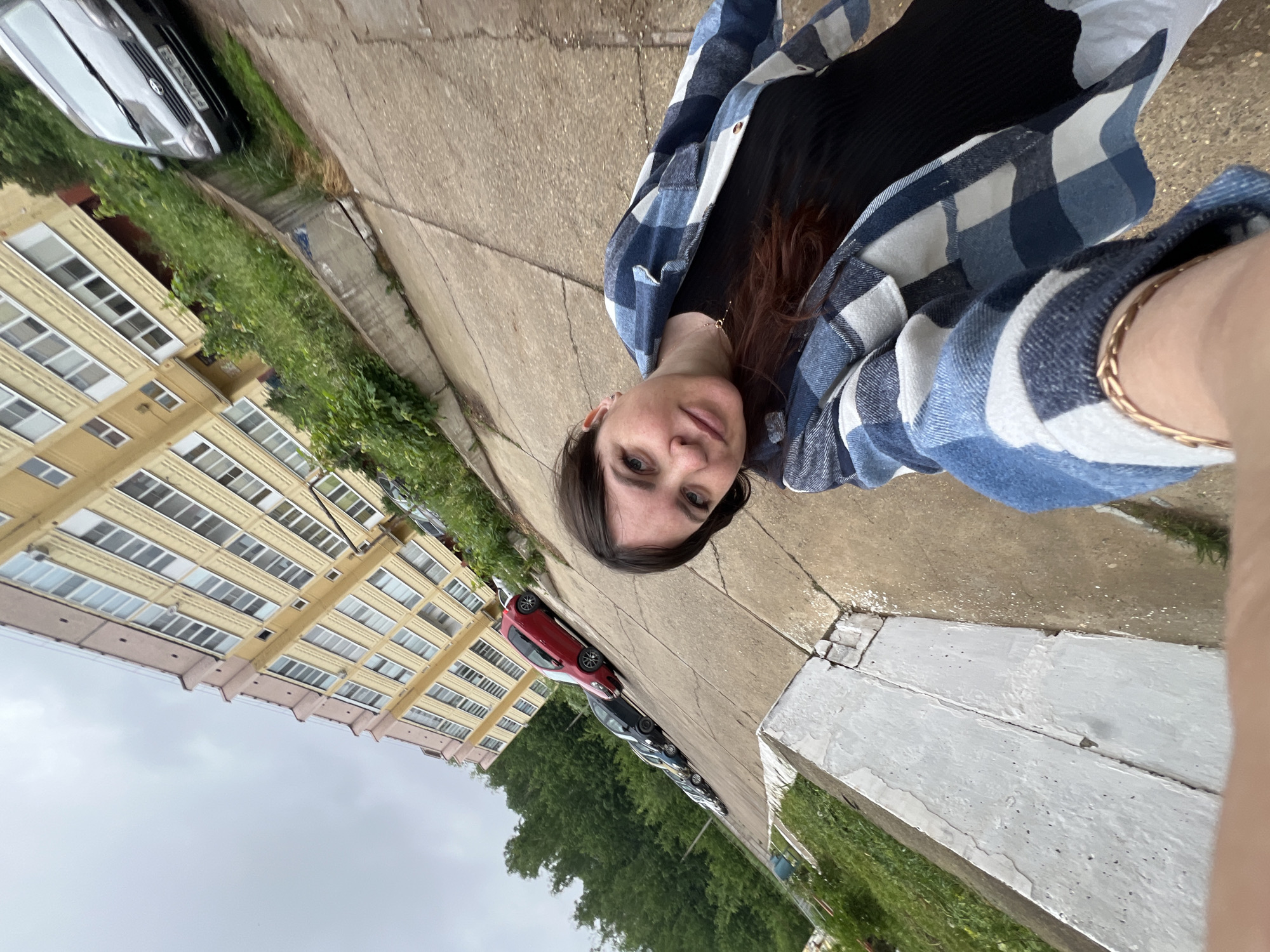A photograph depicts a woman holding a camera at arm's length, presumably taking a selfie. She is wearing a black top and a blue and white plaid flannel shirt. A gold bracelet adorns her wrist. The background features a city street scene. A multi-story apartment building with yellow facade is visible on the left. Parked cars line the street, including a red car. Green trees and other vegetation are visible. The sky is overcast. The photograph seems to be taken in Noviy, Russia.