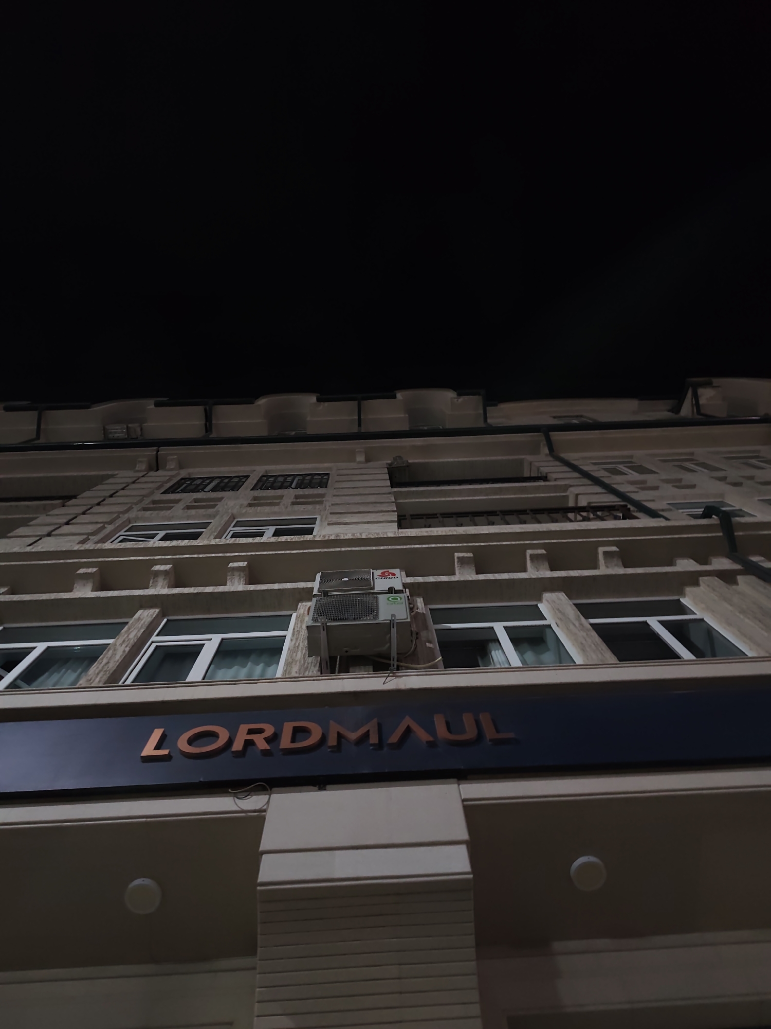 A low-angle exterior shot captures a building facade at night in Fergana City, Uzbekistan. The building is light beige with architectural details resembling stacked blocks and protruding ledges. Multiple windows with white frames are visible on several floors, some with sheer white curtains. An outdoor air conditioning unit is mounted to the facade. Below the windows, a dark blue sign displays the name "LORDMAUL" in orange, three-dimensional lettering. The sky above is dark and unobscured.