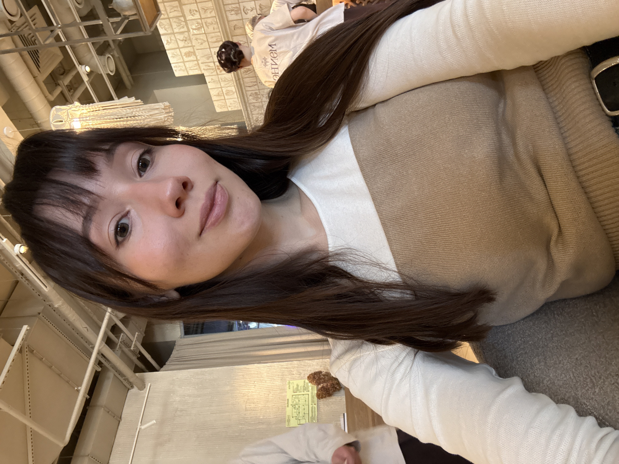 A selfie shot depicts a young woman with shoulder-length dark brown hair and bangs. She's wearing a cream-colored long-sleeved shirt layered under a beige sweater. The background reveals an interior setting with white-painted walls and ceiling. Visible elements include industrial-style ceiling fixtures, suggestive of a cafe or restaurant. A person with dark hair wearing a white shirt is visible in the background. Based on architectural details in the background, the location could be in Kazan, Russia, but confirmation is needed to confirm this statement.