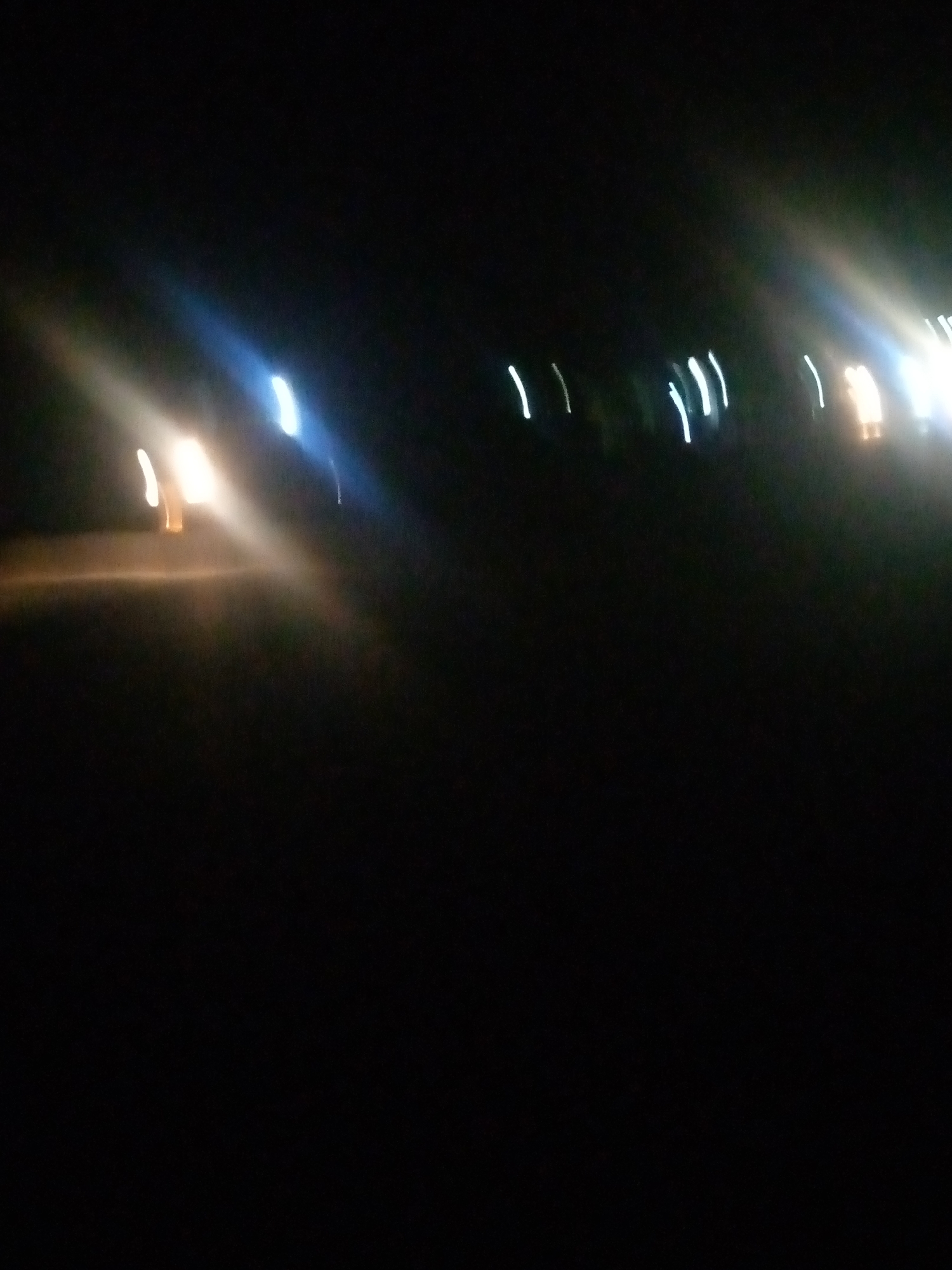 The image captures a low-light scene, likely at night, depicting streaks of light that suggest motion or a long exposure photograph. In the foreground, a bright, elongated streak of yellowish-white light is visible, possibly from headlights of a vehicle. To the right and further back, several vertical white streaks are scattered across the frame, which could represent streetlights or taillights. The background is predominantly dark, with some faint glows and blurs indicating distant lights. The overall impression is one of movement in a dimly lit urban environment. The scene does not provide enough detail to definitively identify a specific location within Jalingo, Nigeria, but the presence of vehicle lights and streetlights is consistent with a road or thoroughfare in any city. No people or specific structures are clearly discernible.