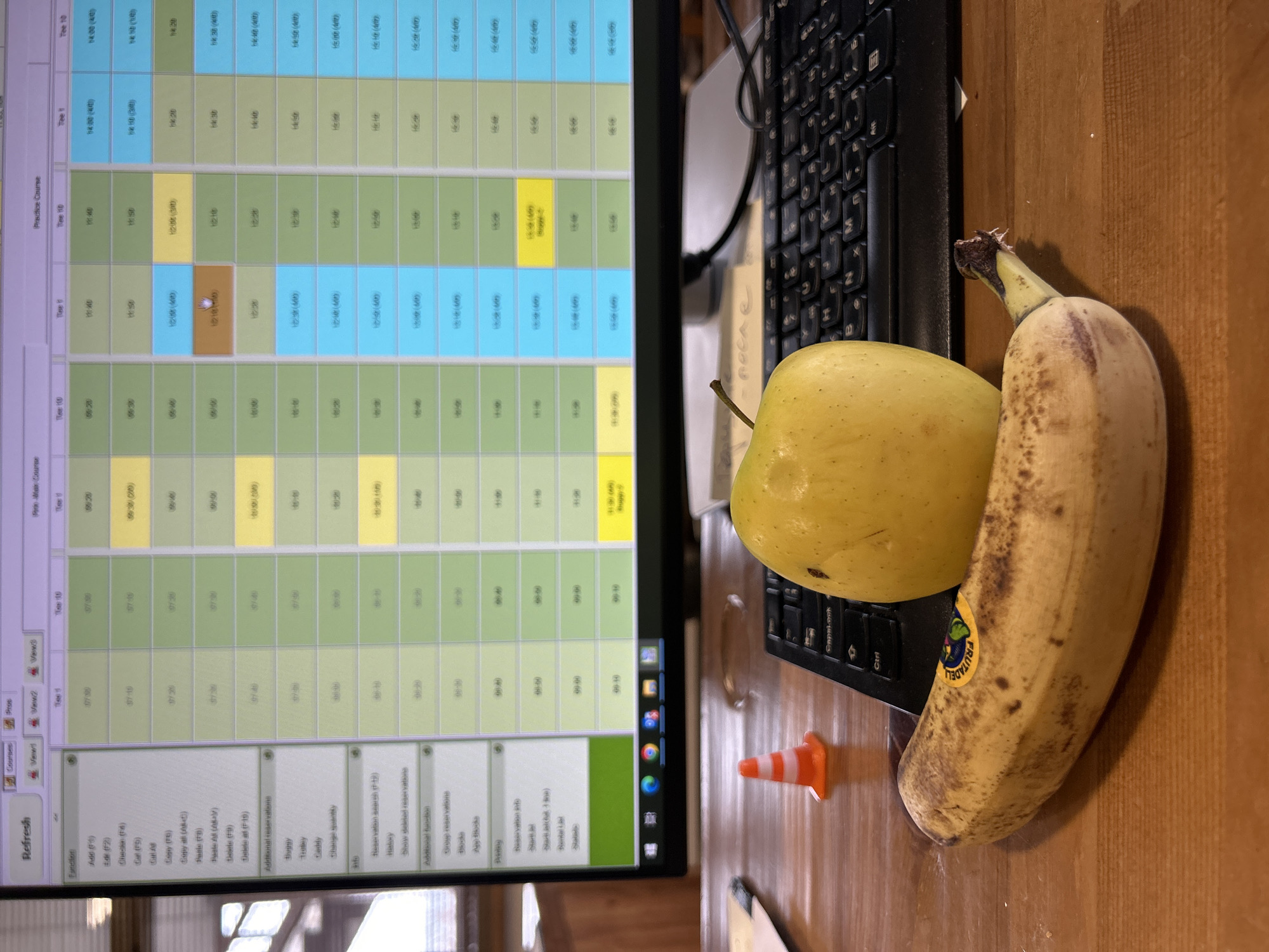 A cluttered desk scene featuring a computer monitor displaying a program with a schedule, a black keyboard, an apple, a banana, and a small orange traffic cone. The monitor is angled slightly to the left, showing a software interface for scheduling, possibly golf tee times. The apple is yellow with a stem, and the banana is yellow with some brown spots and a sticker. The traffic cone is small and orange. The location of the image is not related to Razlog, Bulgaria. The lighting appears to be indoor and natural. There are no people visible in the frame.