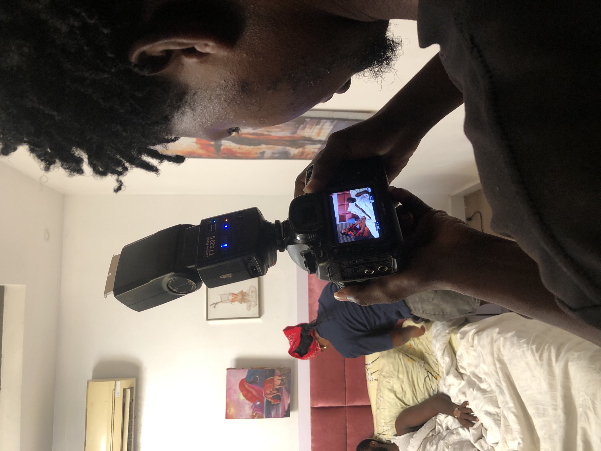 A man with dreadlocks is holding a camera with a flash in a bedroom in the city of Unknown, Unknown. The camera's LCD screen shows an image of people in what appears to be a photograph. In the room, a person wearing a red bandana is kneeling on a bed, while another person lies on the bed, covered by a white sheet. Paintings are hung on the white walls, and an air conditioning unit is mounted on the wall. The lighting is mixed, with both natural and artificial light sources present.