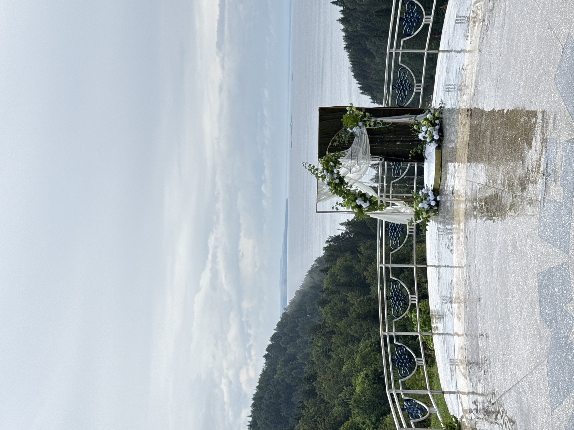 The image depicts an outdoor wedding ceremony setup on a balcony overlooking a body of water and a forested hillside. The balcony floor is wet and reflective, suggesting recent rain. A white railing with decorative blue elements borders the balcony. In the center, a wedding arch is adorned with white and green flowers and draped with sheer white fabric. Behind the arch, a dark backdrop stands. The background features a vast expanse of water, likely a river or lake, leading to a distant shoreline with a densely forested hill. The sky is overcast with clouds. The location is identified as the city of Galevo, Russia.