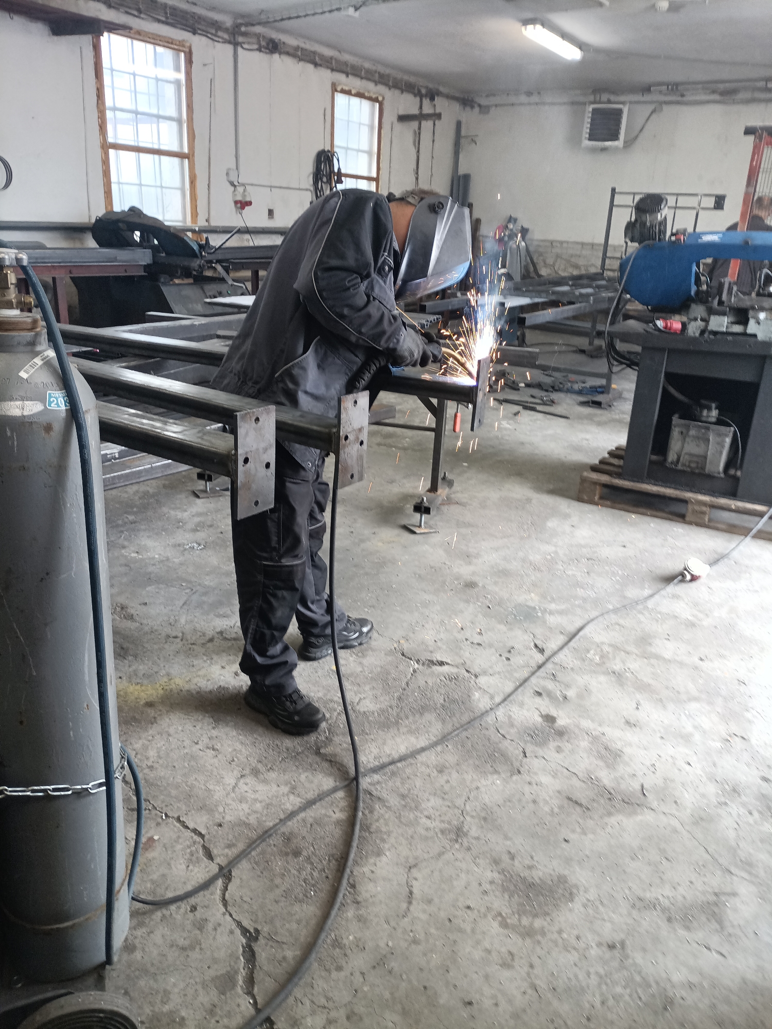 The image depicts a working industrial workshop in Dalovy, Czechia, where a person is actively engaged in welding. The primary subject is an individual, likely male, dressed in dark work coveralls or jacket and pants, wearing protective gloves and a welding helmet. Bright sparks emanate from the point where they are welding a metal structure, which appears to be a frame or assembly made of square steel tubing, set up on adjustable metal stands.
To the left, a large, tall gray gas cylinder, possibly for shielding gas used in welding, is visible. It is secured by a chain and has hoses connected to it. Visible text on the cylinder includes "203" and other partial numbers like "2021" and possibly "MESO".
The workshop floor is concrete, exhibiting cracks, stains, and scattered metal debris, indicative of a busy work environment. In the background, various pieces of equipment are present, including a blue horizontal metal-cutting bandsaw on a black stand, other metal tables, and machinery. A wooden pallet sits beneath one of the machines. The walls are painted a light color, featuring several multi-paned windows with wooden frames, suggesting an older building. Fluorescent lighting fixtures are mounted on the ceiling, supplementing natural light. Electrical conduits, a red industrial power outlet, and a white rectangular heater or ventilation unit are also visible on the walls. The scene conveys a functional, well-used, and practical workspace during daylight hours.