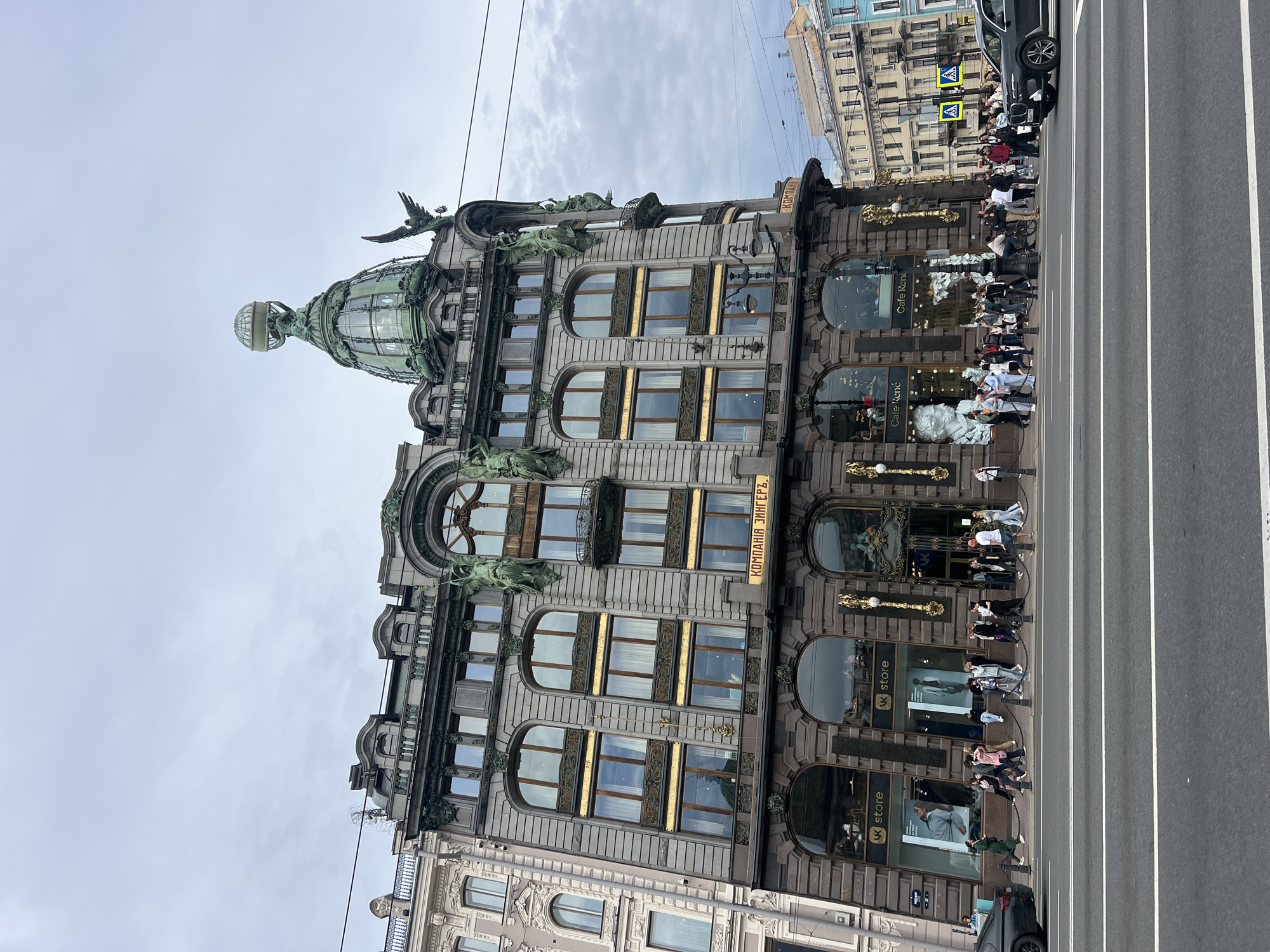 The image shows the Singer House, also known as the Dom Knigi, located at Nevsky Prospekt, 28, Saint Petersburg, Russia. The imposing building features a distinctive green dome topped with a glass globe and eagle sculptures adorning its facade. It has multiple stories with large windows, some featuring gold trim, and ornate architectural details. The ground floor has storefronts, including "VK store" and "Cafe René." A crowd of pedestrians is gathered on the sidewalk in front of the building. A street with cars and traffic lights is in the foreground, with the overcast sky providing a diffused light.