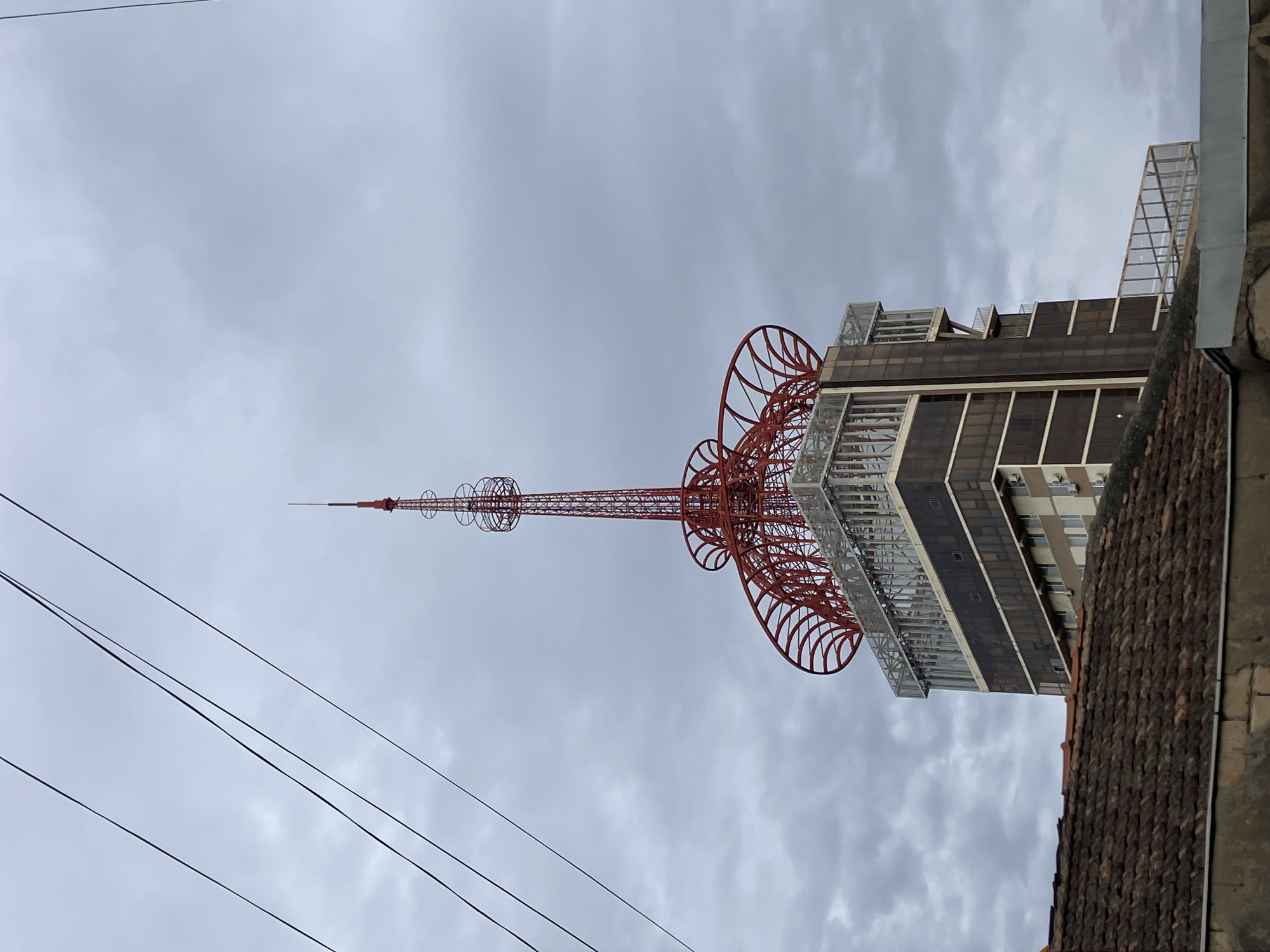 A striking red telecommunication tower dominates the scene, rising prominently from an adjacent modern building against a cloudy, overcast sky. The tower, characteristic of the Tbilisi TV Tower in Georgia, features a slender spire at its peak and an elaborate, multi-tiered structure composed of intricate, spiraling lattice-work and spherical elements, giving it a distinctive and artistic silhouette. Its vibrant red color contrasts sharply with the muted tones of the sky and the building.
The building it stands beside is a multi-story structure with dark, reflective windows and a contemporary design. Its upper levels feature angular, metallic-framed architectural details, possibly observation decks or technical installations, which partially integrate with the base of the tower.
In the foreground, several dark power lines stretch diagonally across the lower left portion of the image, suggesting an urban setting. To the lower right, a section of an old, terracotta-tiled rooftop and a glimpse of a stone wall are visible, hinting at traditional architecture in the immediate vicinity, contrasting with the modern infrastructure.
The sky is filled with soft, grey clouds, indicating an overcast day. There are no people visible, and no specific activity is taking place; the scene captures a static architectural landmark. No discernible text is visible within the image.