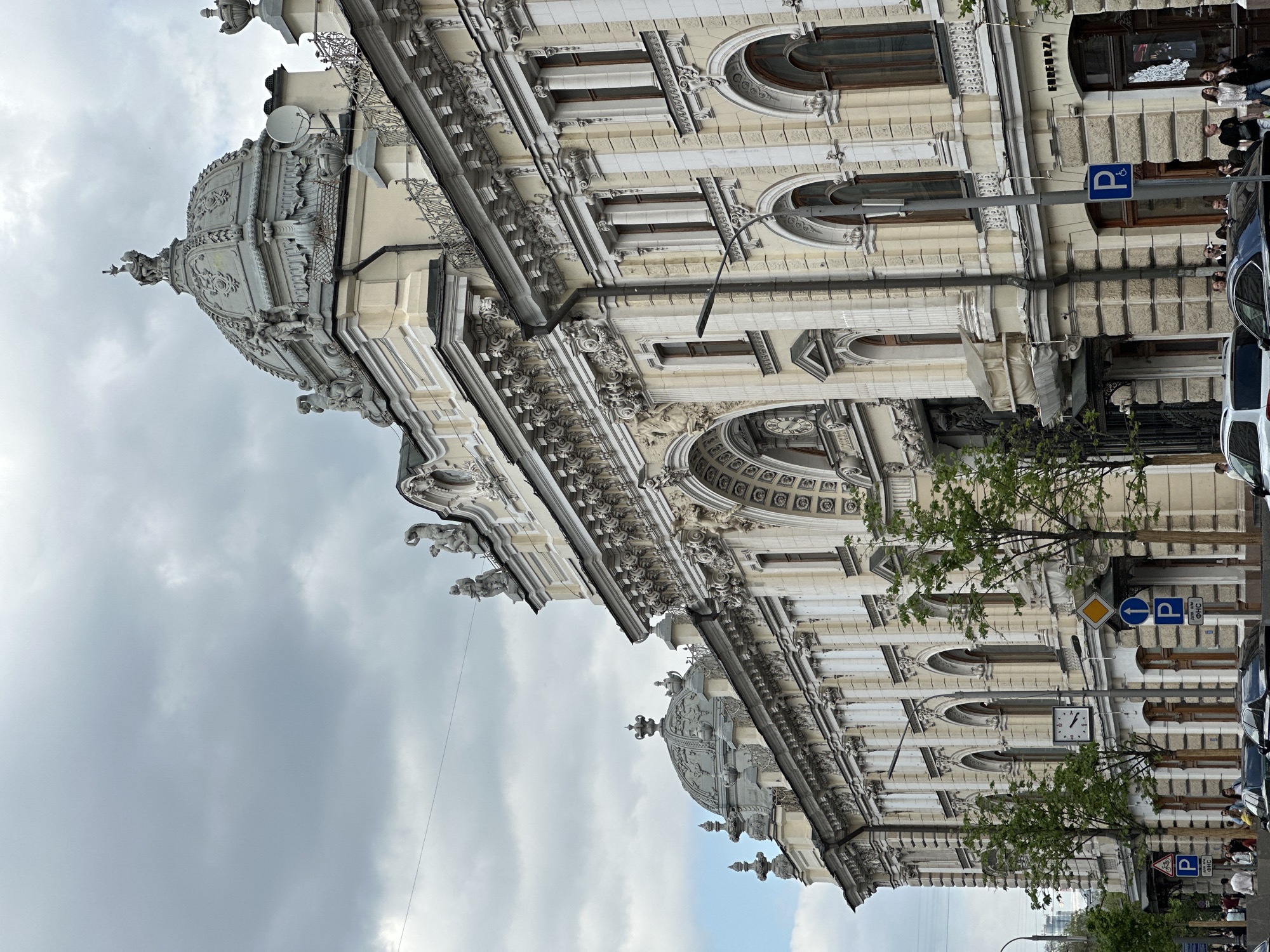 The image captures the facade of a grand, ornate building located in Moscow, Russia. The architectural style is elaborate, featuring detailed carvings, domes, arched windows, and columns. The building is likely an historical structure.
The scene is at street level, with trees and street signs visible. There are parked cars and pedestrians present, indicating an active urban environment. The sky overhead is overcast. The image is vertically oriented.