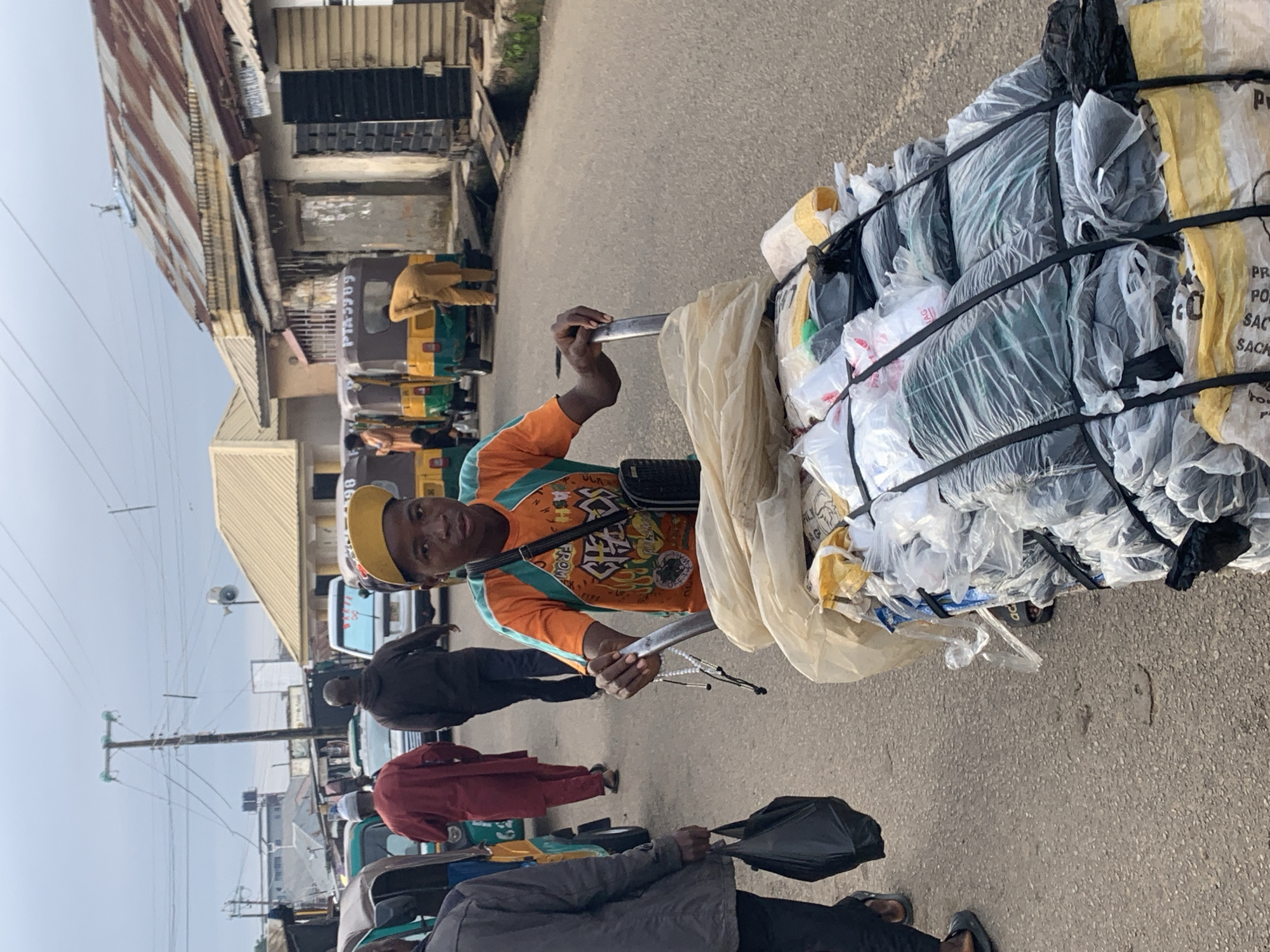 A young Black male wearing a yellow baseball cap and an orange and teal shirt is pushing a cart loaded with goods wrapped in plastic. The cart is made of metal with black straps holding the load in place. He appears to be on a street in Jos, Nigeria, as indicated by the presence of auto-rickshaws, commonly known as "Keke Napep," in the background. Other people are visible in the background, including a person in a red robe and another in a dark jacket walking away from the camera. Buildings with corrugated iron roofs line the street. The sky is overcast. The cart contains a large quantity of items, mostly obscured by clear plastic wrapping.